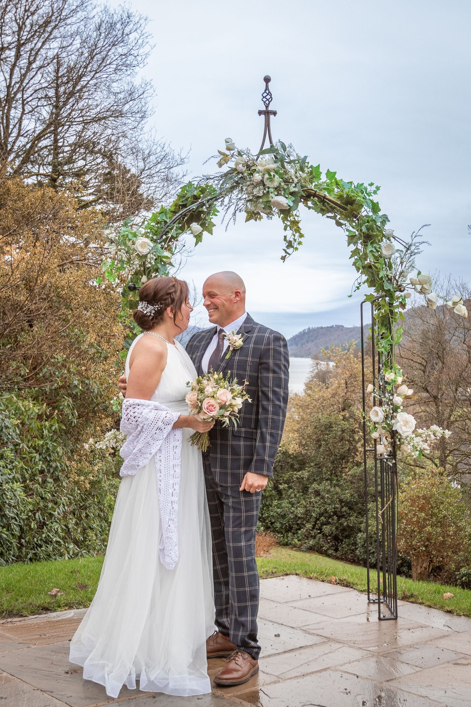Bride and groom stand under floral arch with lake views, exchanging vows outdoors in autumn wedding ceremony.