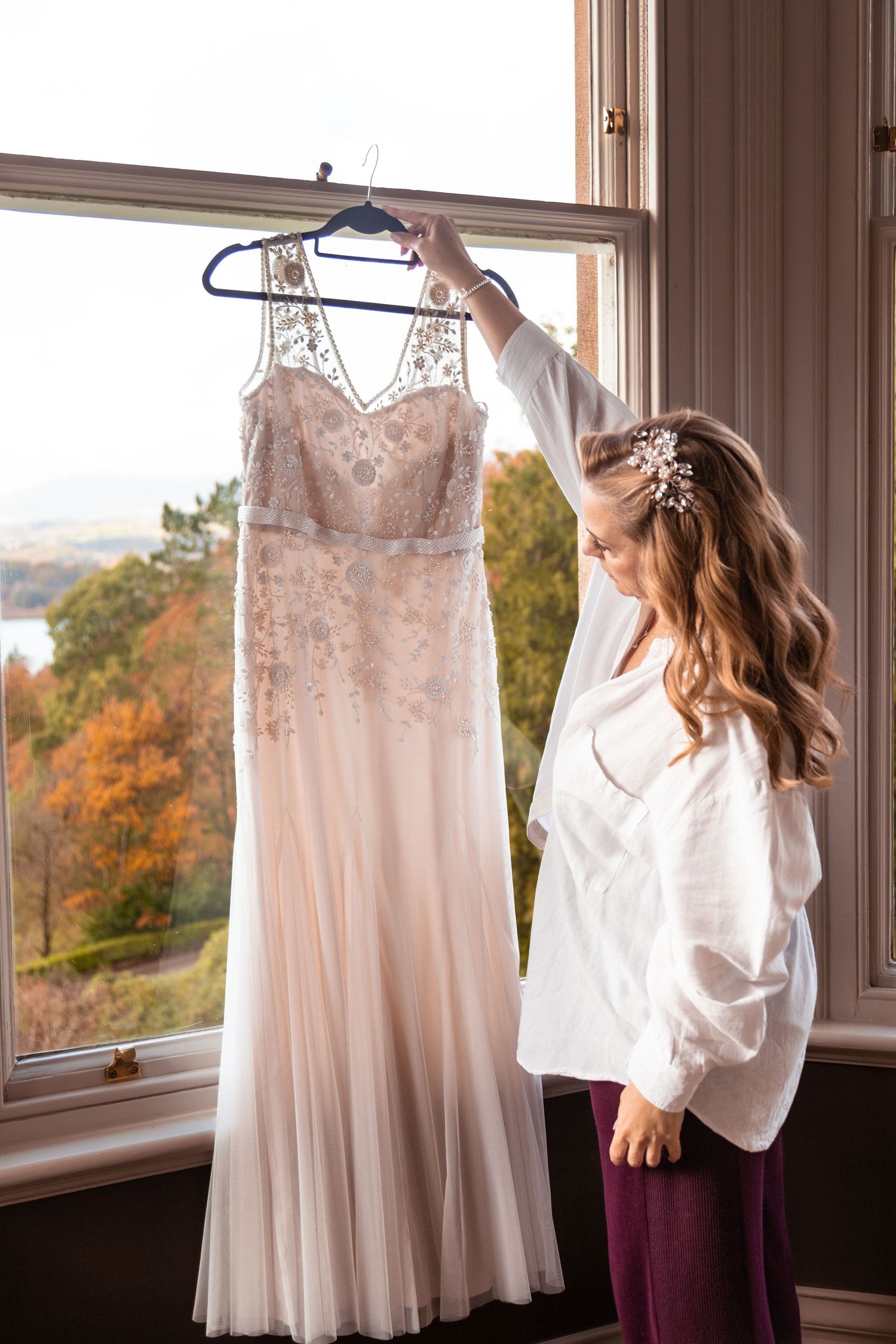 Bride admiring beaded wedding gown hanging by autumn window.