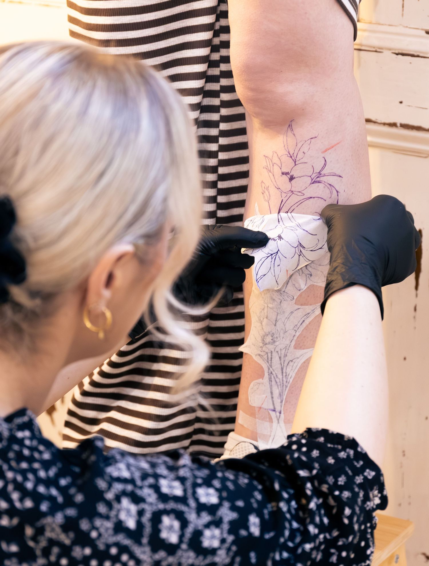 Tattoo artist applying floral stencil transfer to client's leg wearing black gloves.