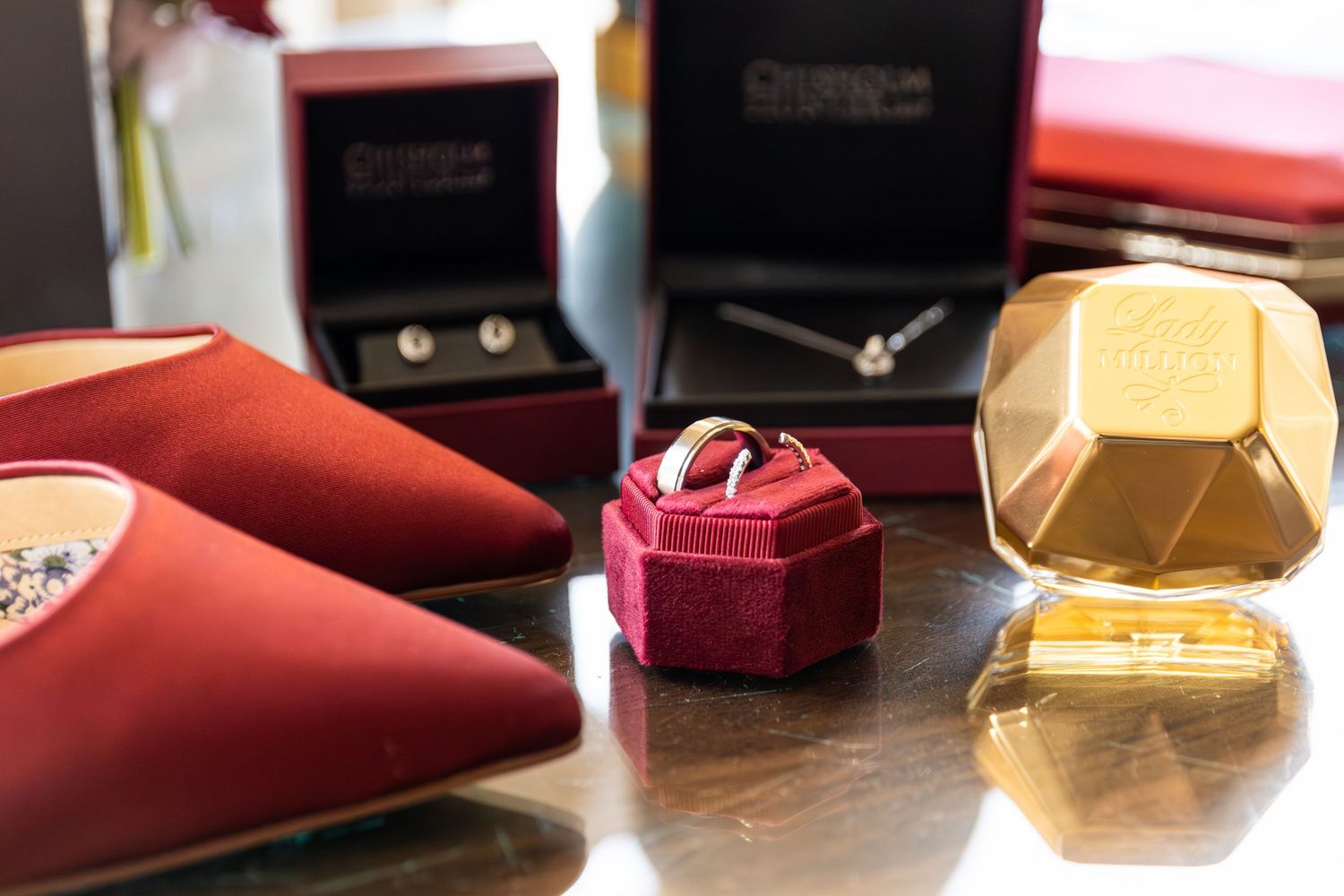 Wedding accessories including red heels, ring box, earrings, and gold perfume bottle on a reflective surface.