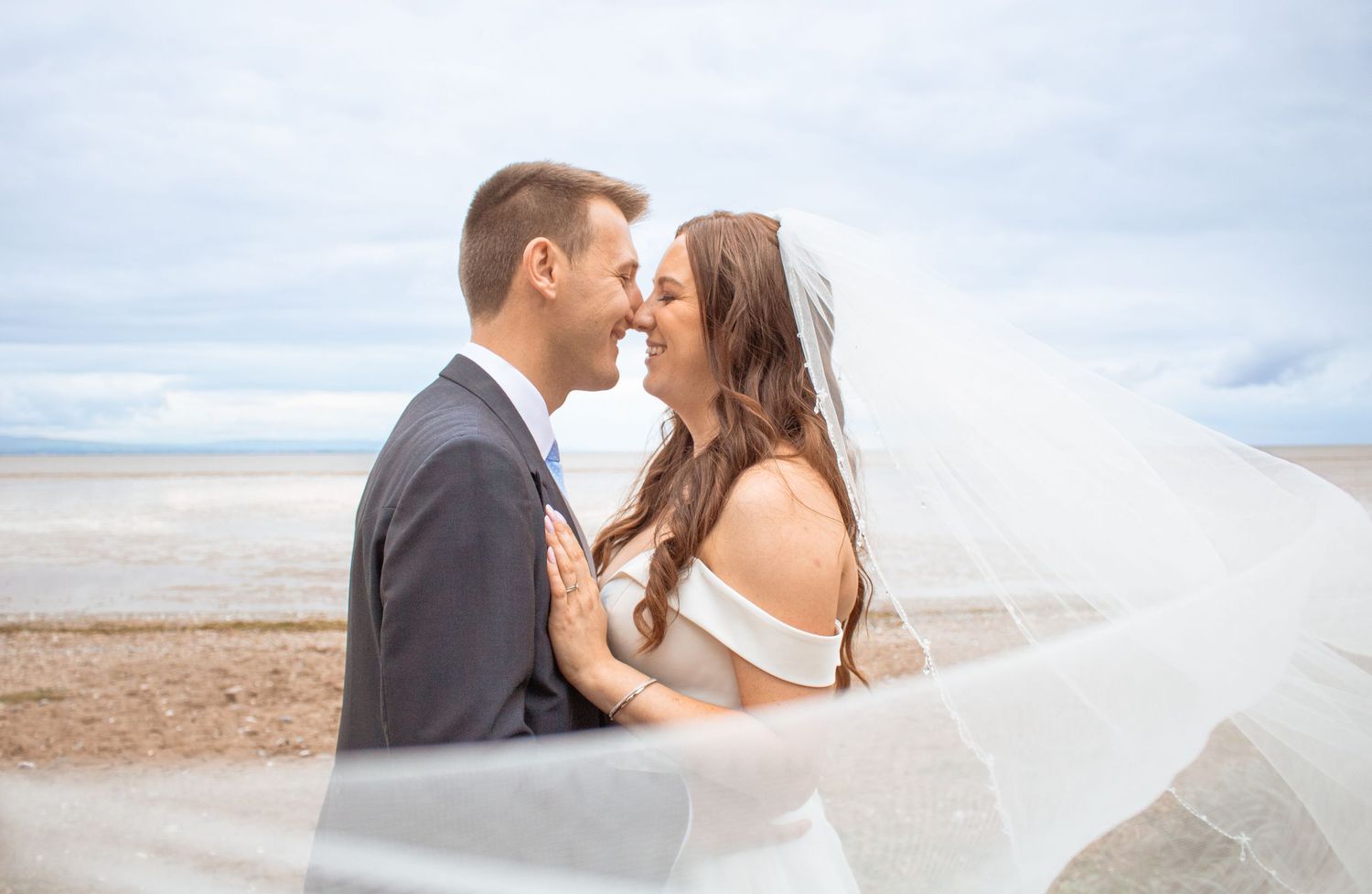 Bride and groom share a romantic moment on a sandy beach with veil flowing in the wind.