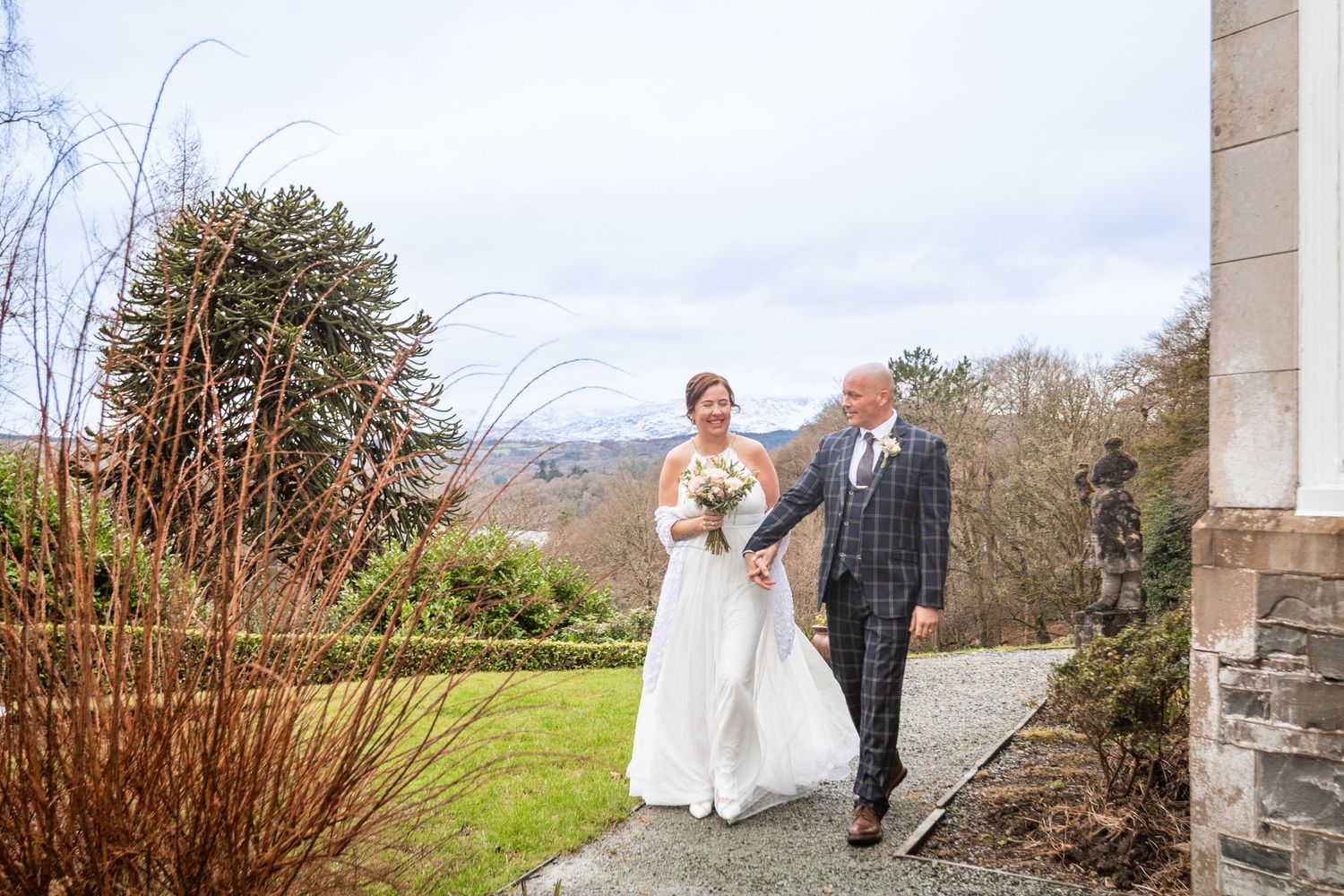 Bride and groom walking together through garden grounds of a historic wedding venue on a cloudy day.