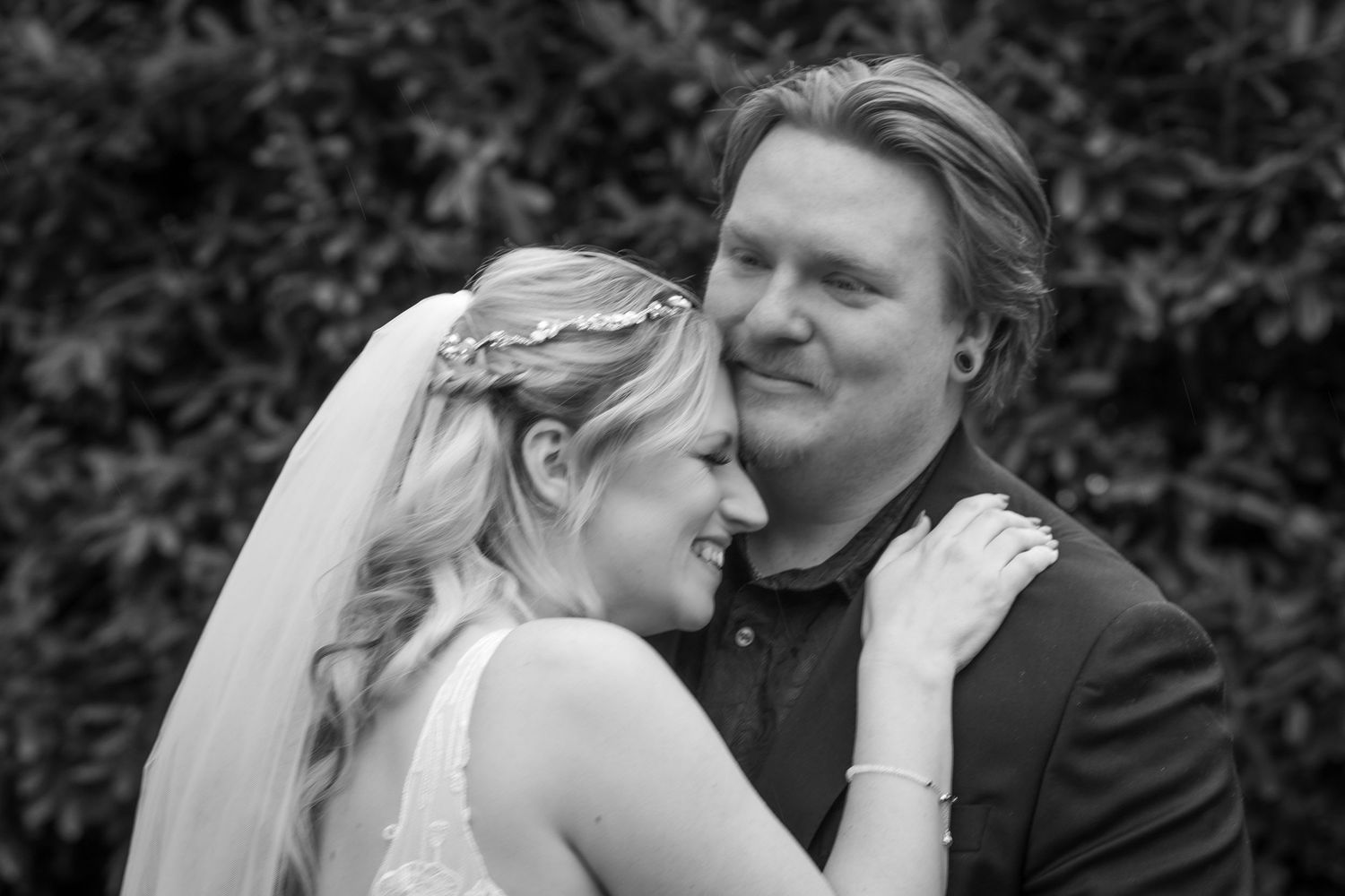 Black and white wedding photo of a smiling bride in veil and groom embracing outdoors near green foliage.