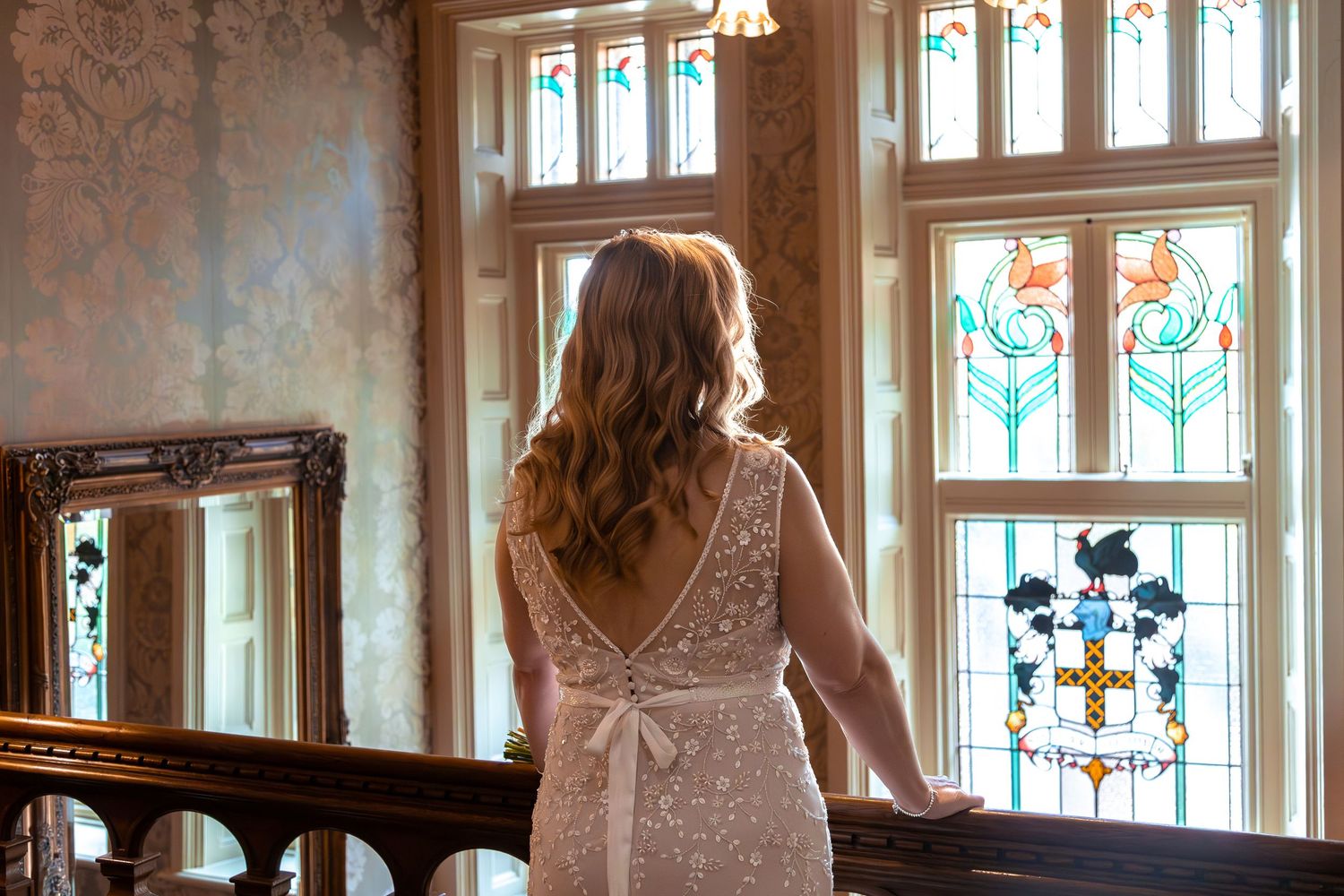 Bride in backless lace wedding gown stands before ornate stained glass windows in elegant historic mansion.