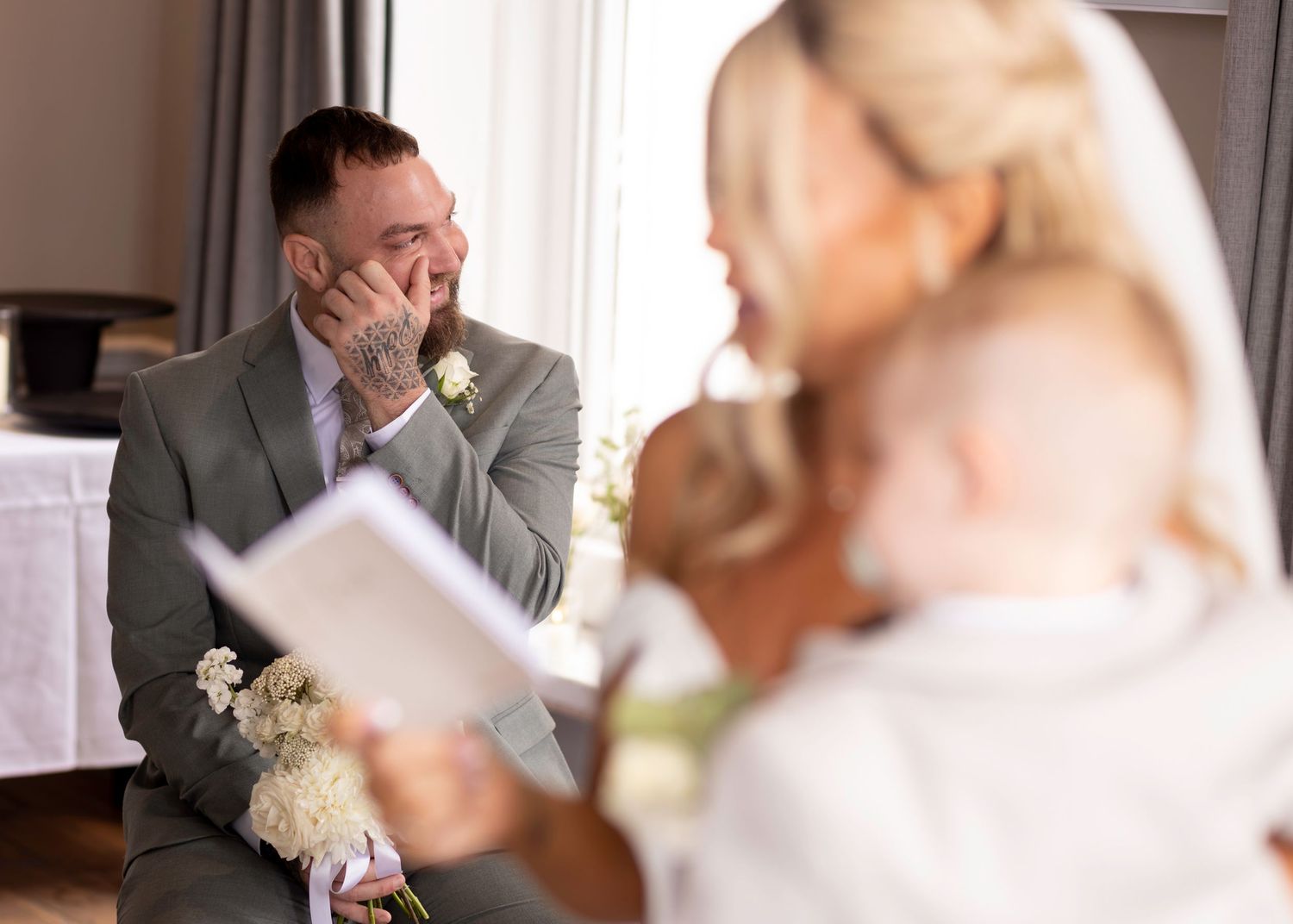 Emotional groom wipes tears as bride reads wedding vows while holding baby during intimate ceremony.