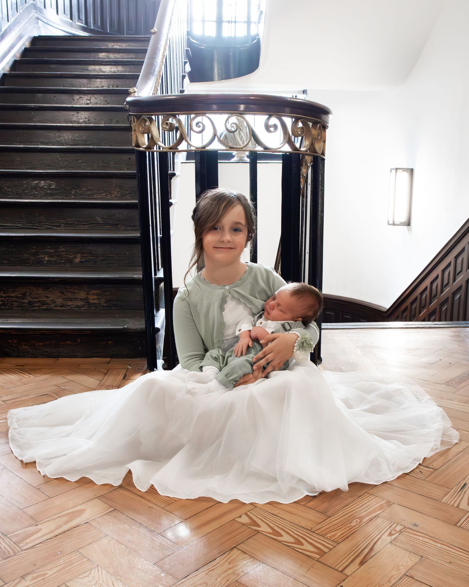 Young girl in white tulle skirt sits on hardwood floor holding newborn baby near elegant staircase.