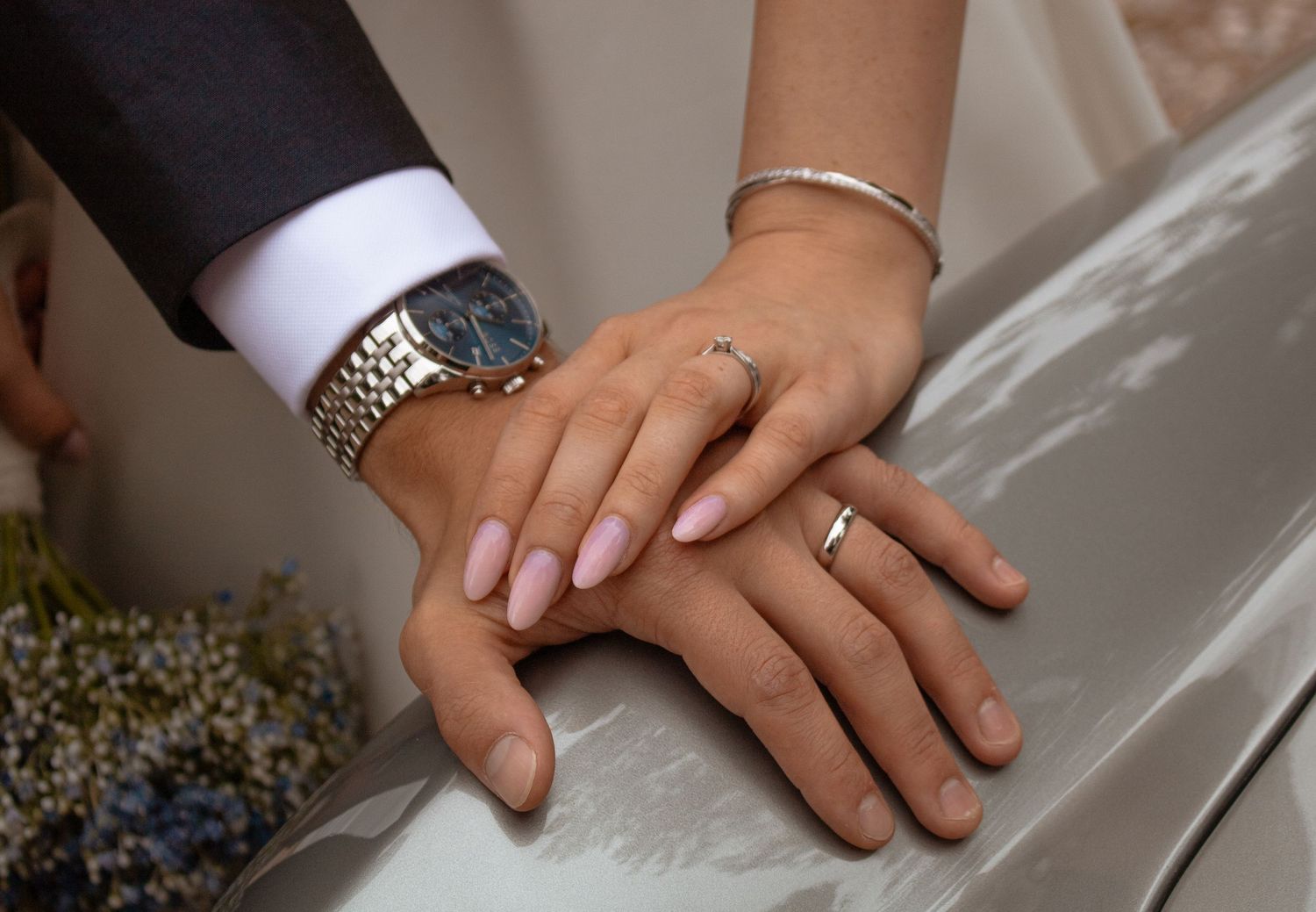 Close-up of newlyweds hands showing wedding rings, bride with pink nails and bracelet, groom wearing gold watch.