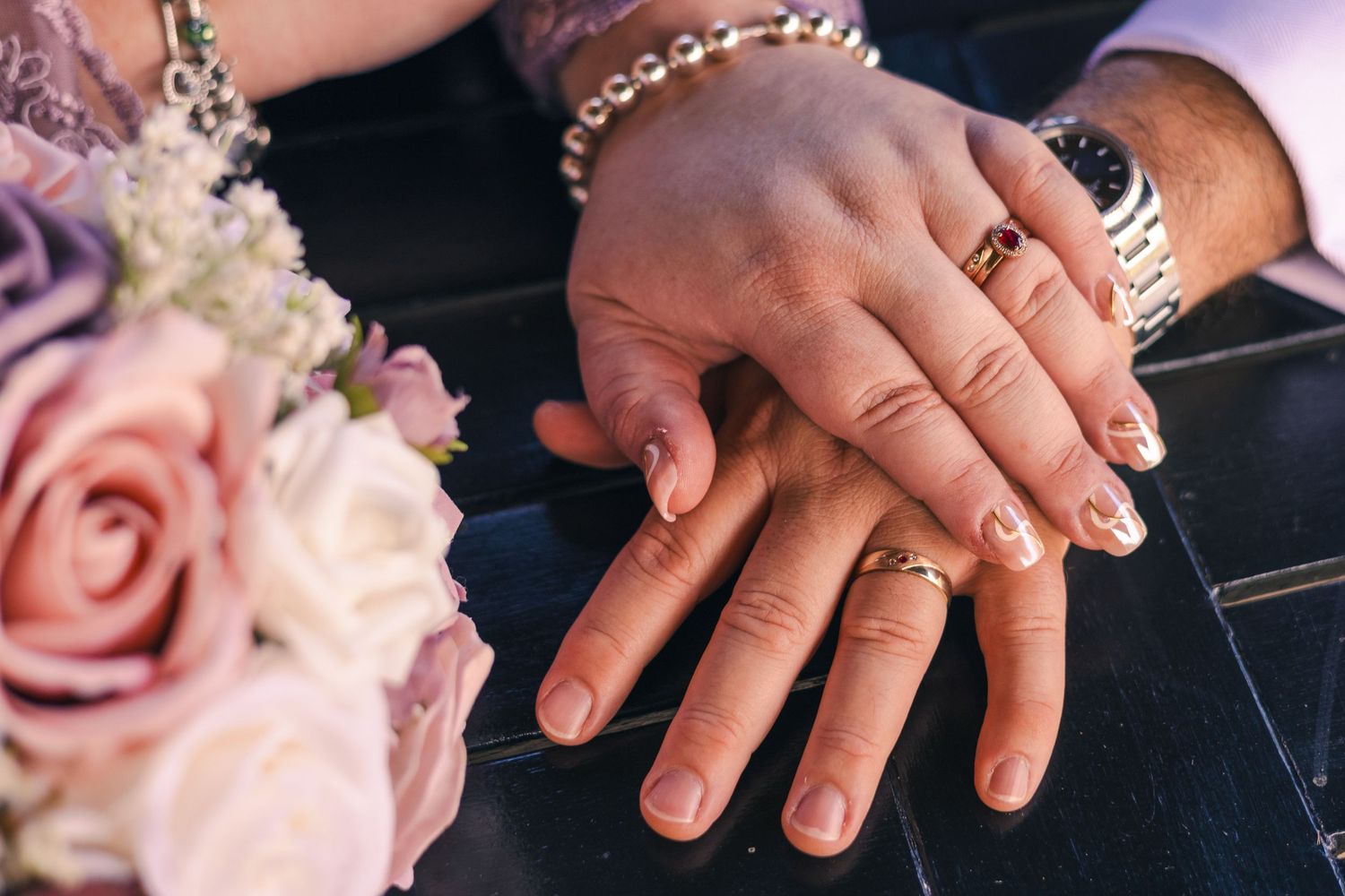 A newlywed couple displays their wedding rings with hands resting near a pink rose bouquet.