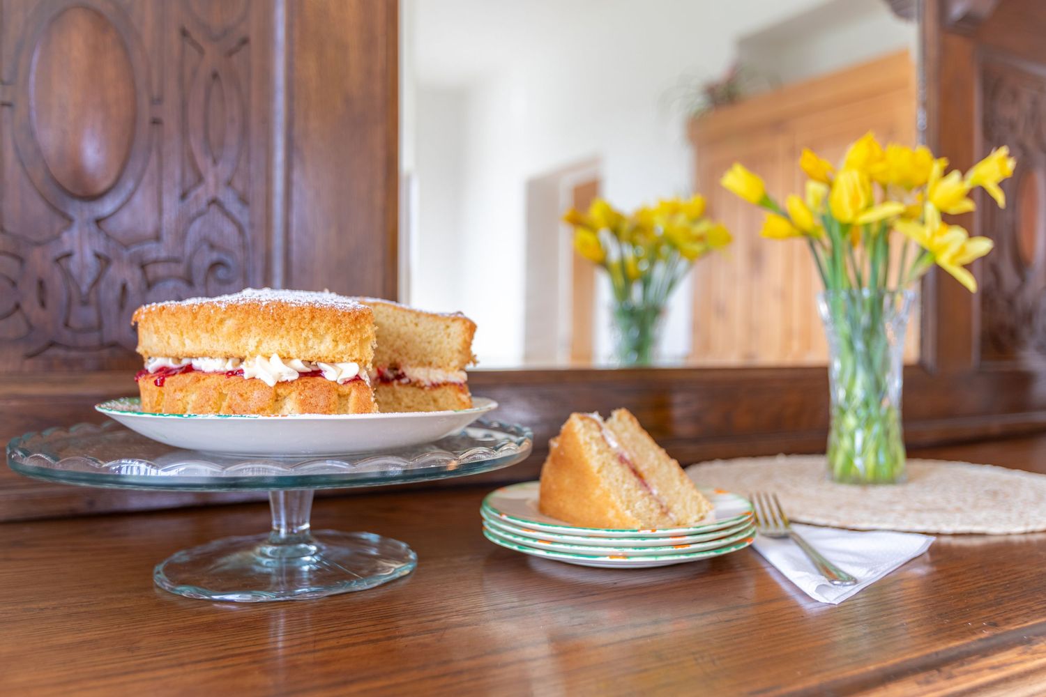 Classic Victoria sponge cake on a glass stand beside fresh yellow daffodils on a wooden sideboard.