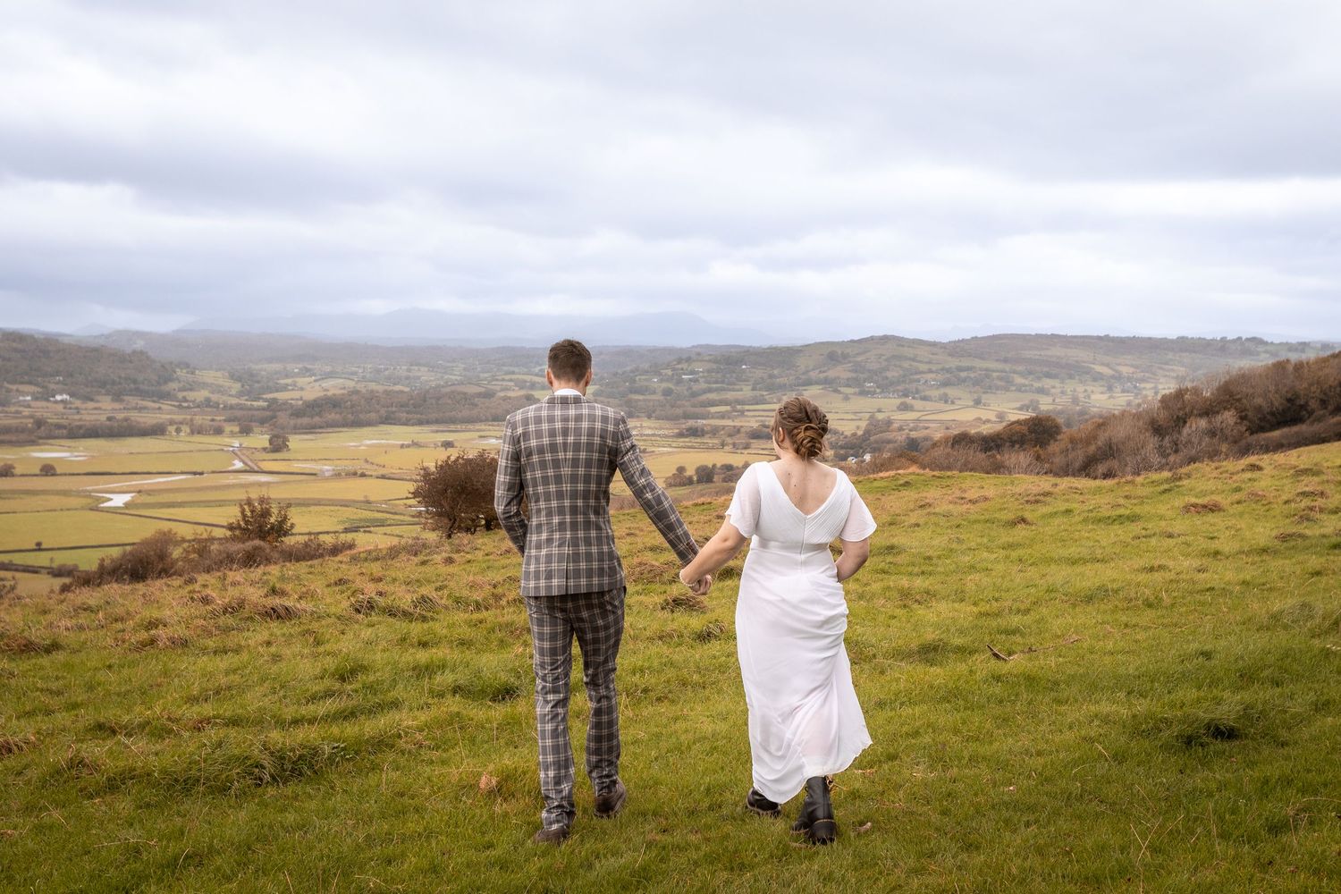 Bride and groom walking hand in hand across green hills with a panoramic valley view.