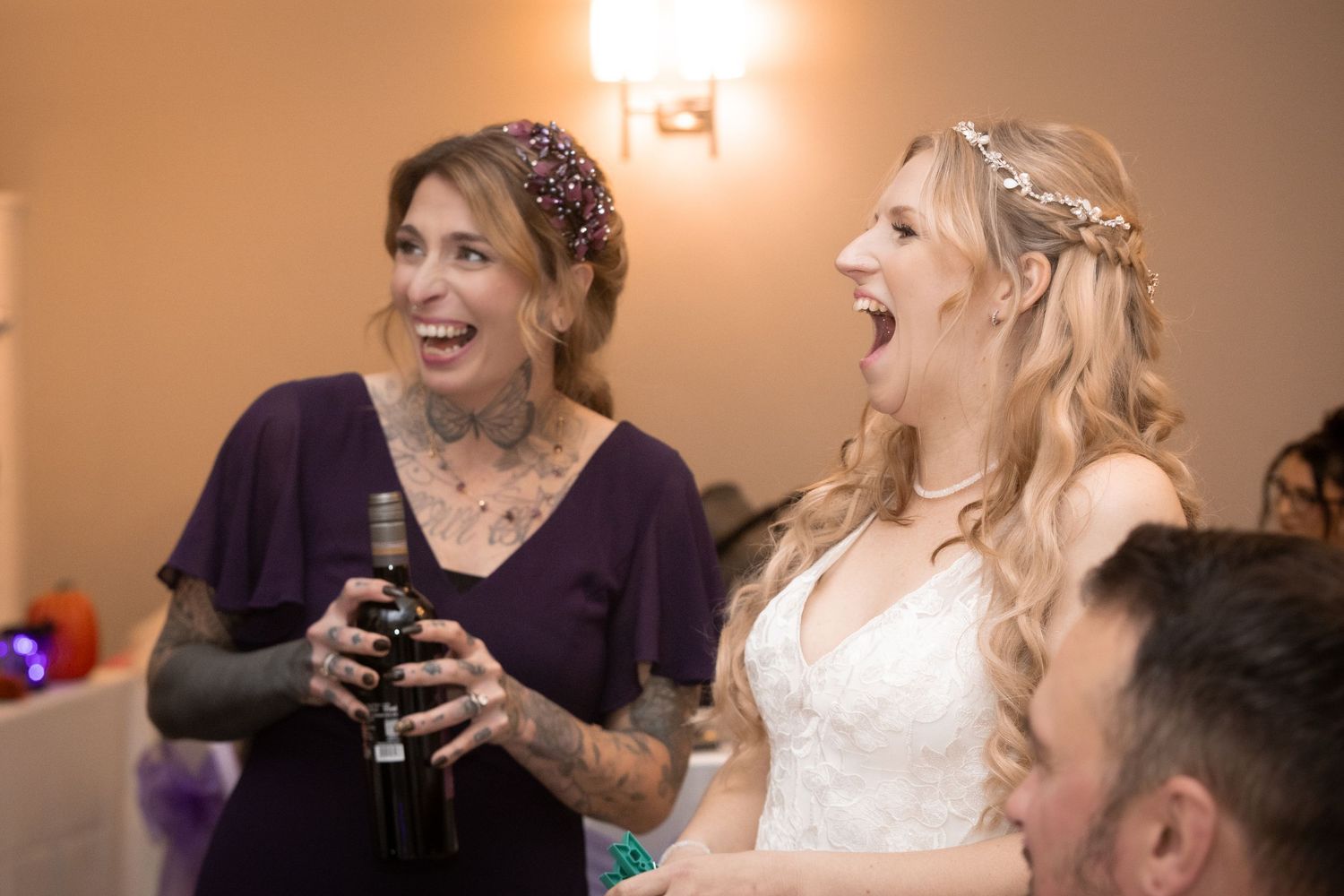 Bride in white lace gown laughing with tattooed woman in purple dress at wedding reception celebration.