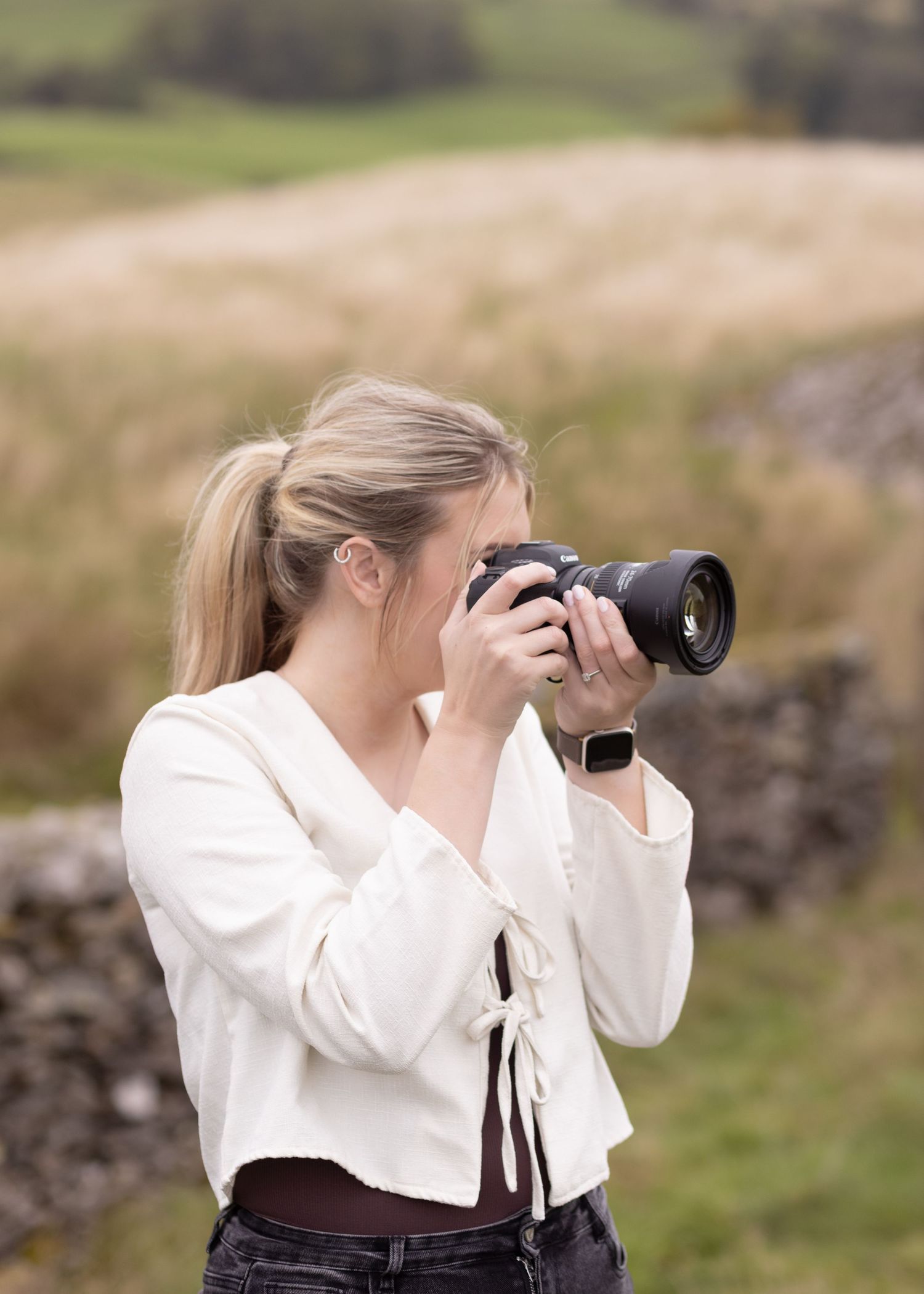 Blonde woman in white top photographing with DSLR camera in rural countryside landscape.