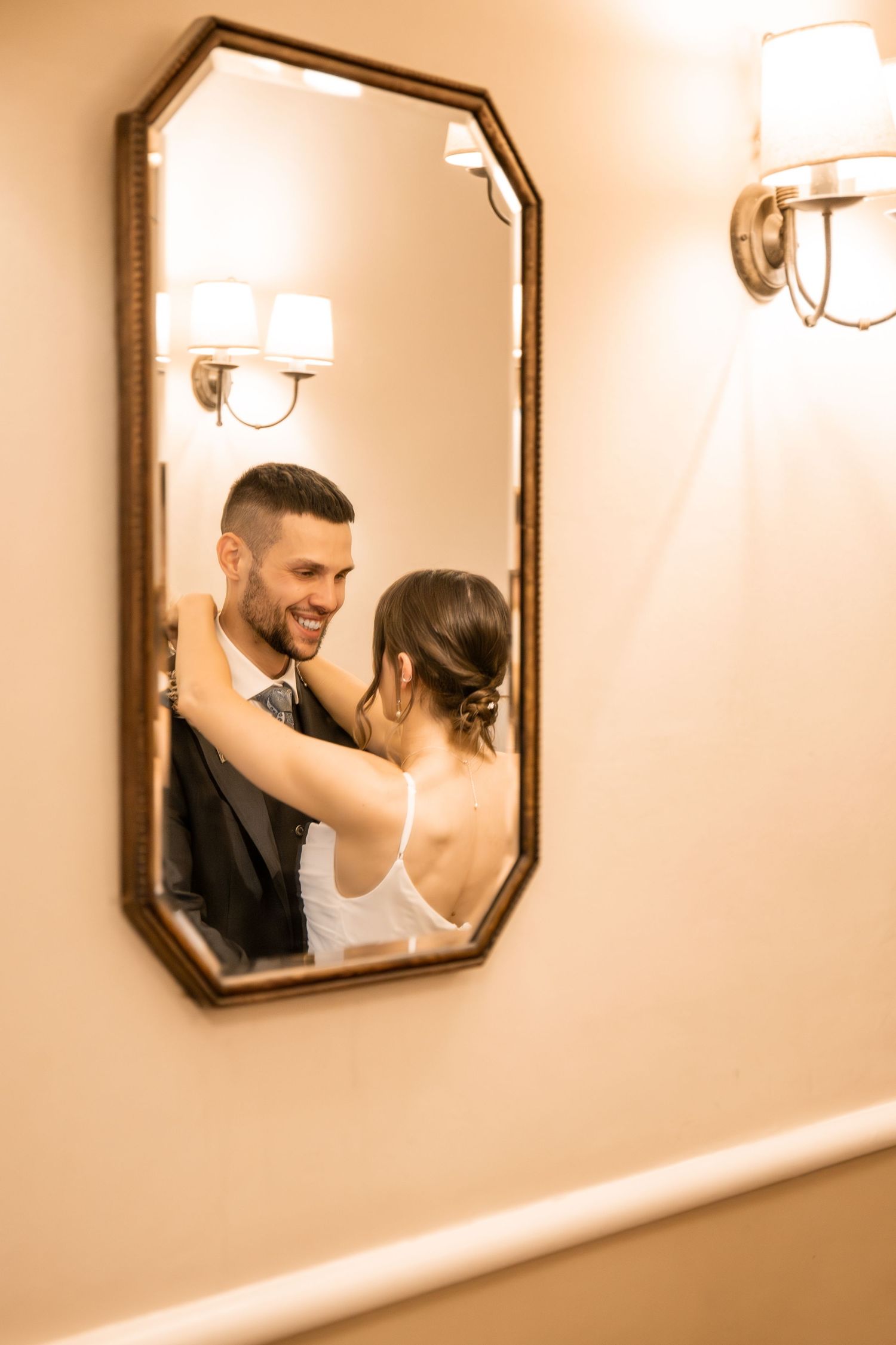 Bride and groom share a romantic embrace reflected in an ornate gold mirror, warmly lit by elegant wall sconces.