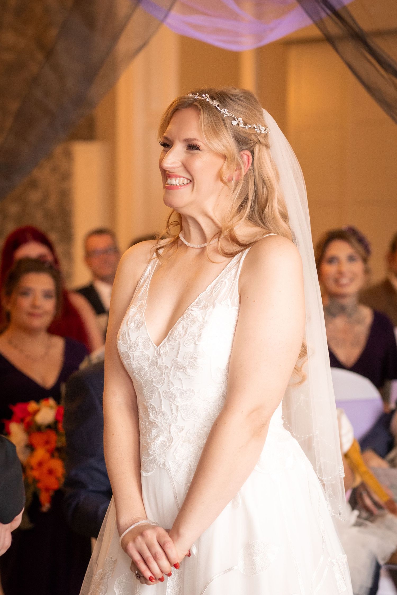 Smiling blonde bride in a lace wedding dress with veil stands at the altar during her ceremony.