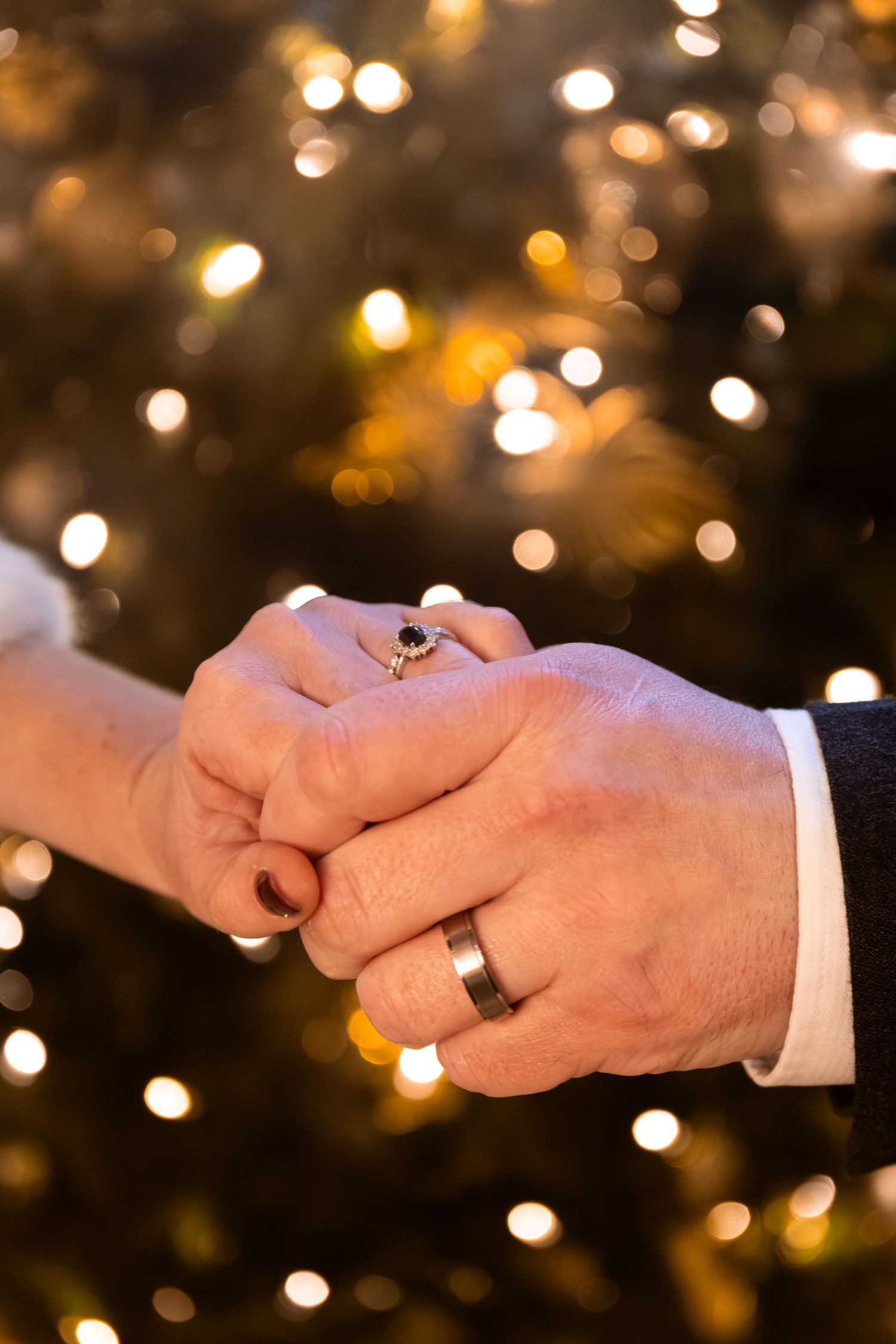 Close-up of newlyweds holding hands showing wedding rings against a bokeh golden lights background.