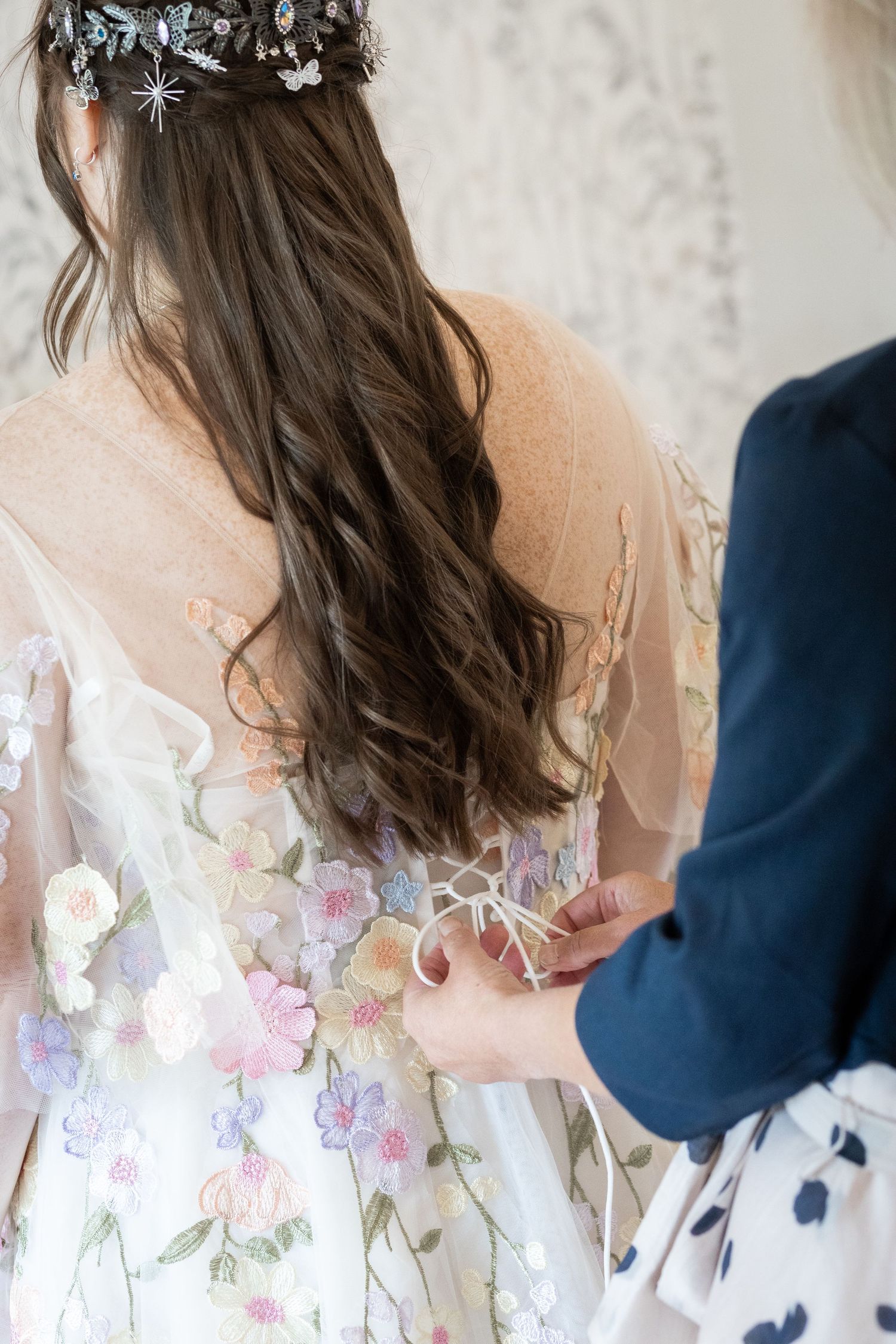 Bride in floral embroidered wedding dress with colorful blooms being laced up, wearing decorative hair crown.