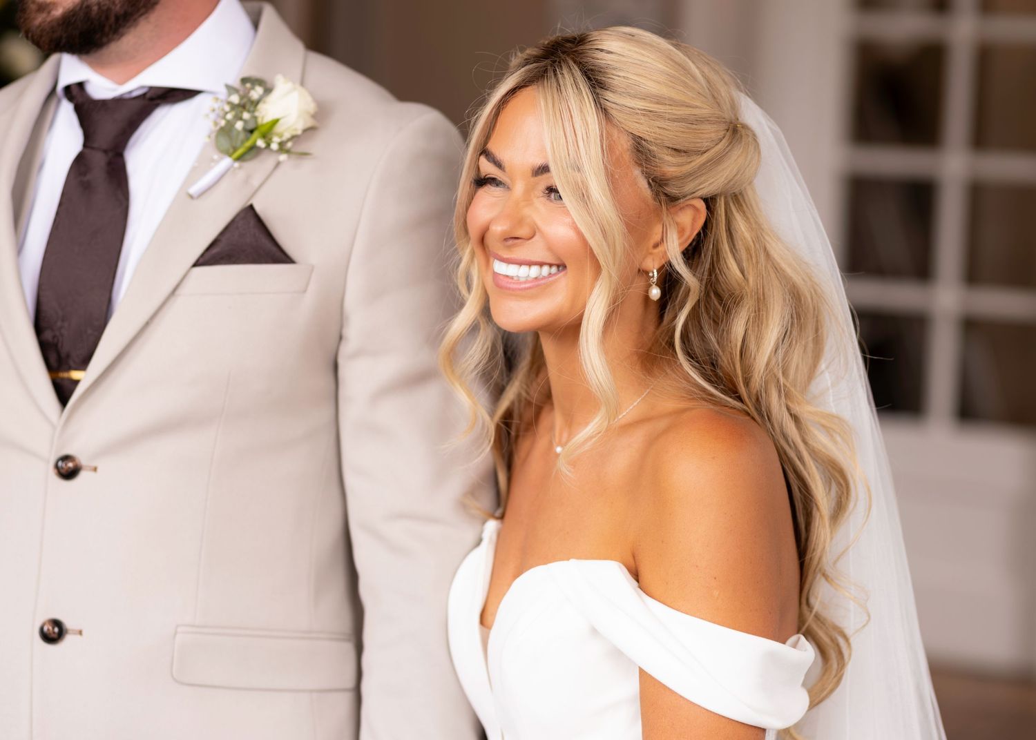 Smiling bride in off-shoulder white gown with flowing veil beside groom in tan suit at wedding.