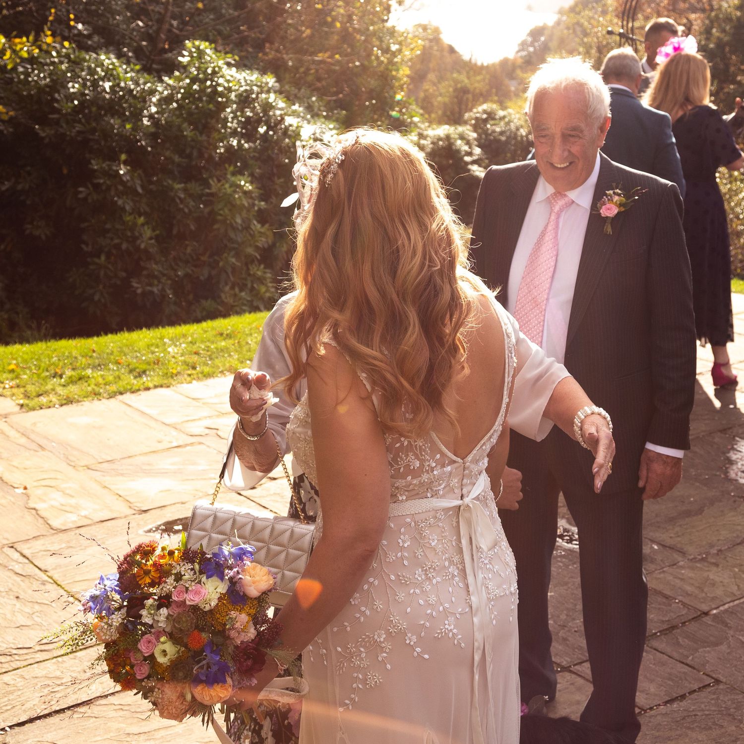 Bride in white gown holding colorful bouquet walks with elderly man in suit on sunny garden patio.