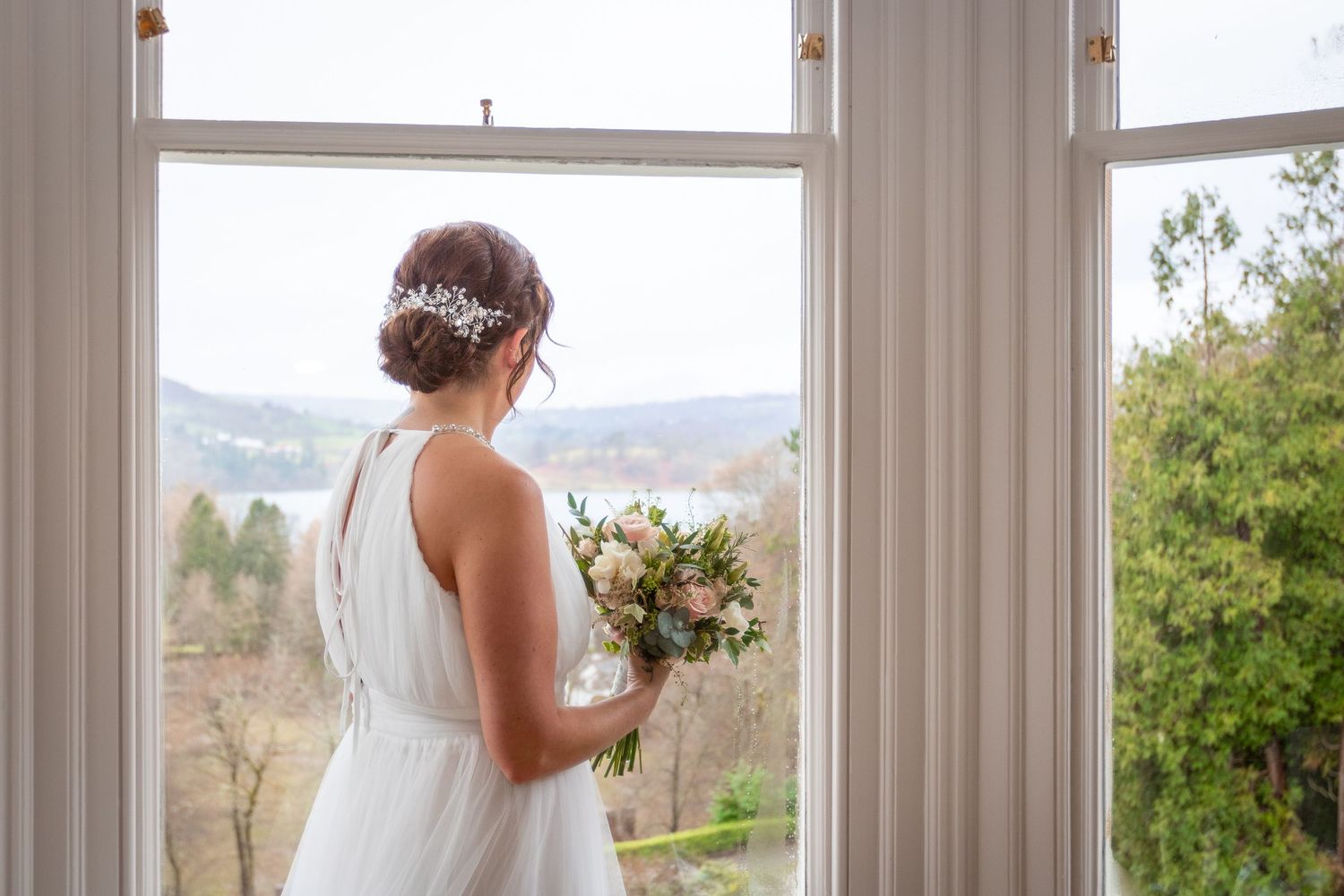 Bride in white dress holding pink bouquet stands by large window with scenic view on her wedding day.