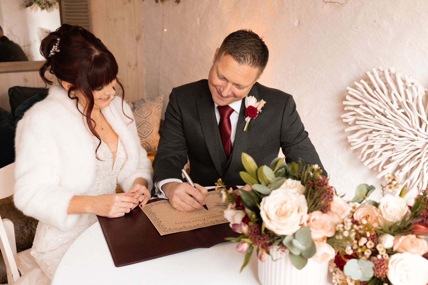 Bride and groom signing wedding registry documents at decorated table with floral arrangement.