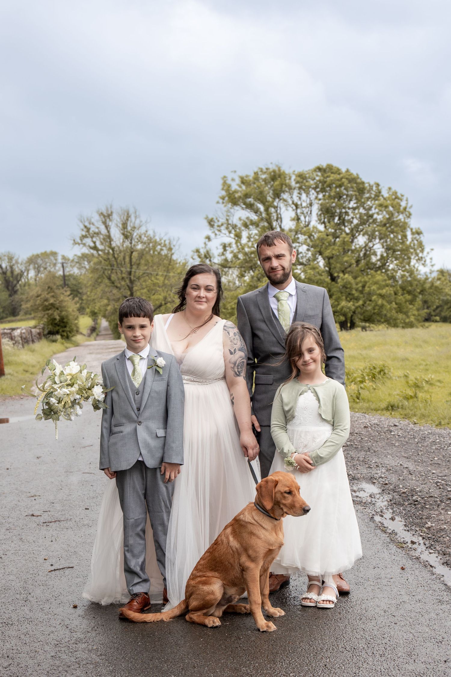 Wedding couple posing with two children and a golden retriever dog on a country road, bride holding white bouquet.