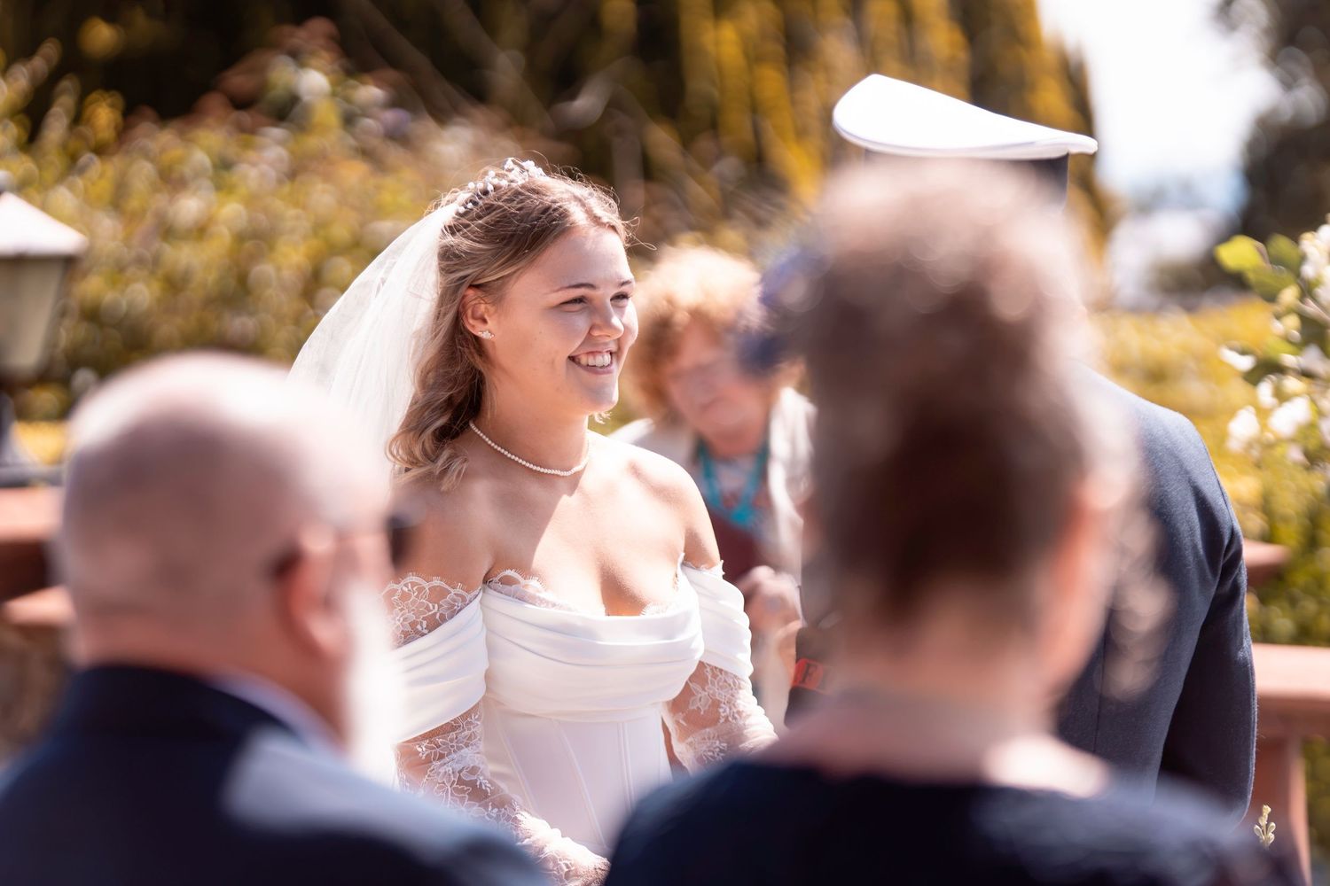 Smiling bride in white off-shoulder gown and veil during outdoor wedding ceremony on a sunny day.