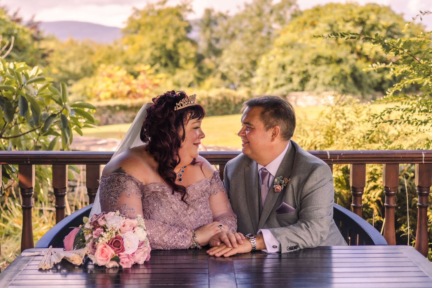 A bride and groom share a romantic moment at an outdoor table surrounded by lush green scenery.