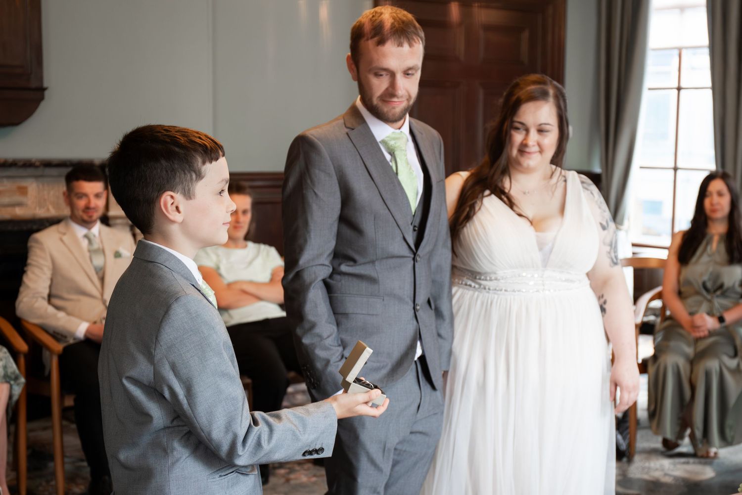 A young boy in a gray suit reads during a wedding ceremony while the bride and groom stand nearby smiling.