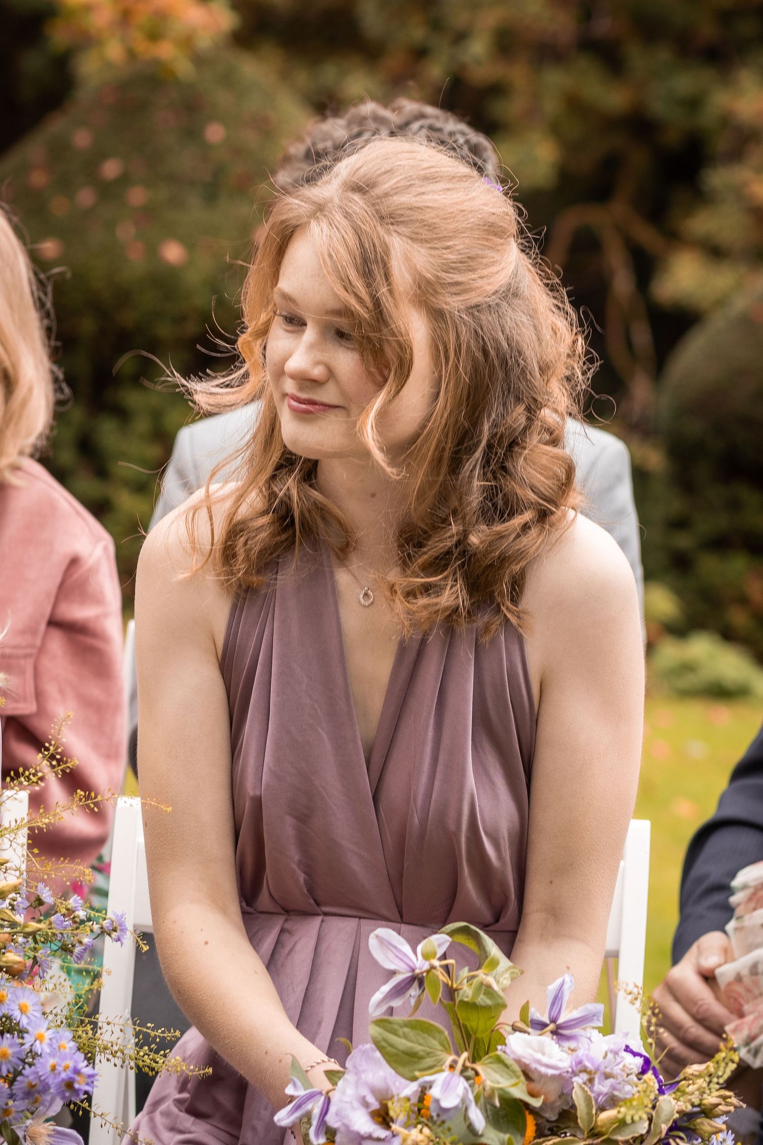 Young woman in mauve bridesmaid dress holding purple flowers smiles at outdoor wedding ceremony.