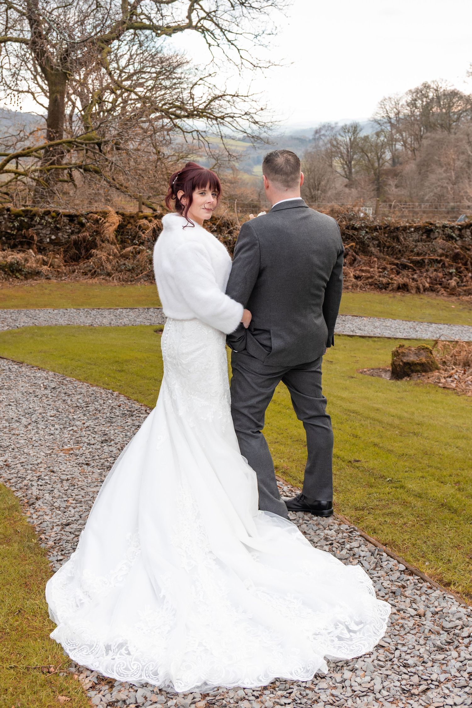 Bride in white lace gown and fur wrap holds groom's hand in winter garden, glancing back at camera.