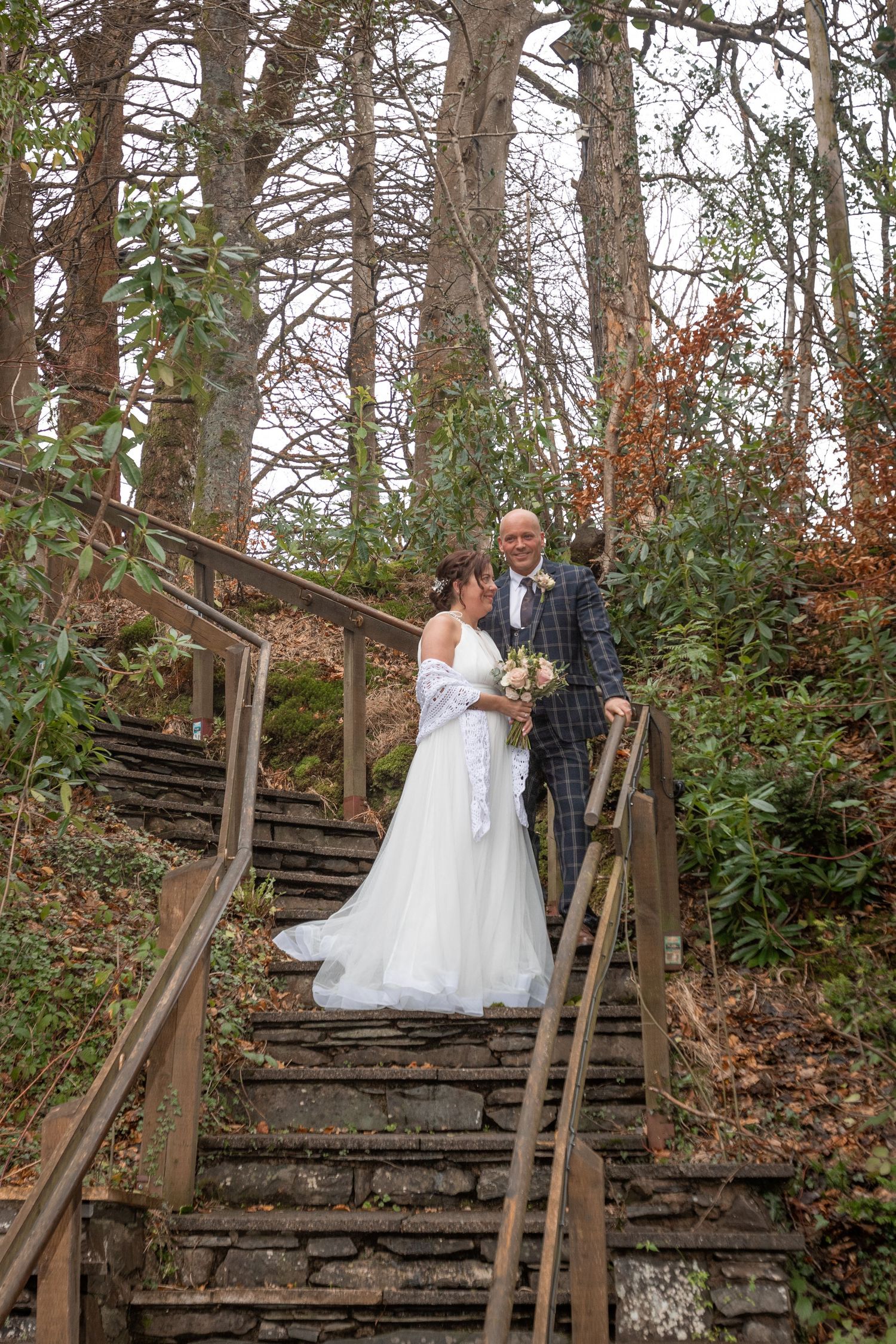 Bride and groom pose on stone steps surrounded by autumn woodland trees, bride holding bouquet in white gown.