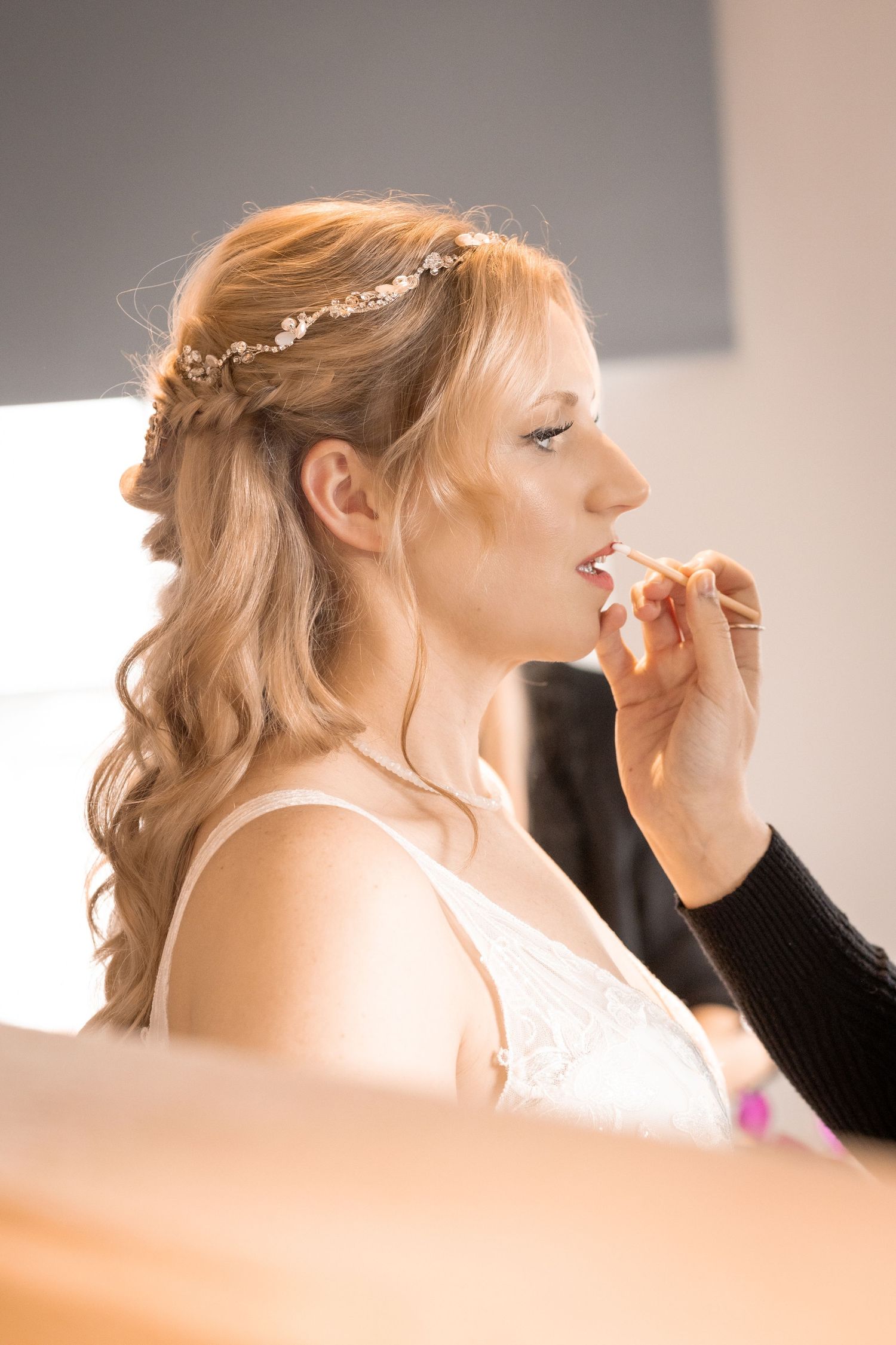 Bride getting lipstick applied by makeup artist before wedding ceremony, wearing floral hair accessory.