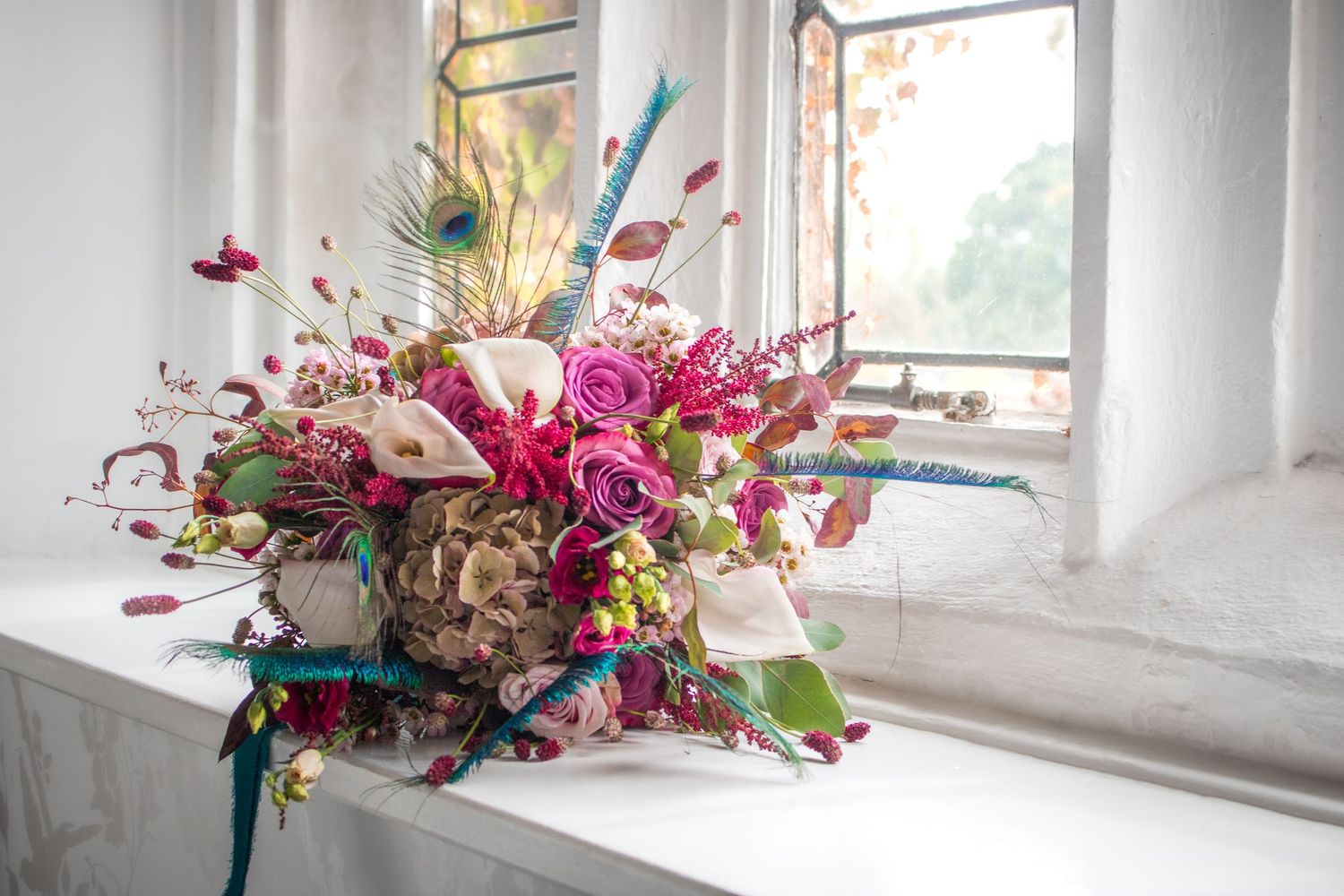 Vibrant colorful wedding bouquet with pink roses, peacock feathers, and mixed blooms on a windowsill.
