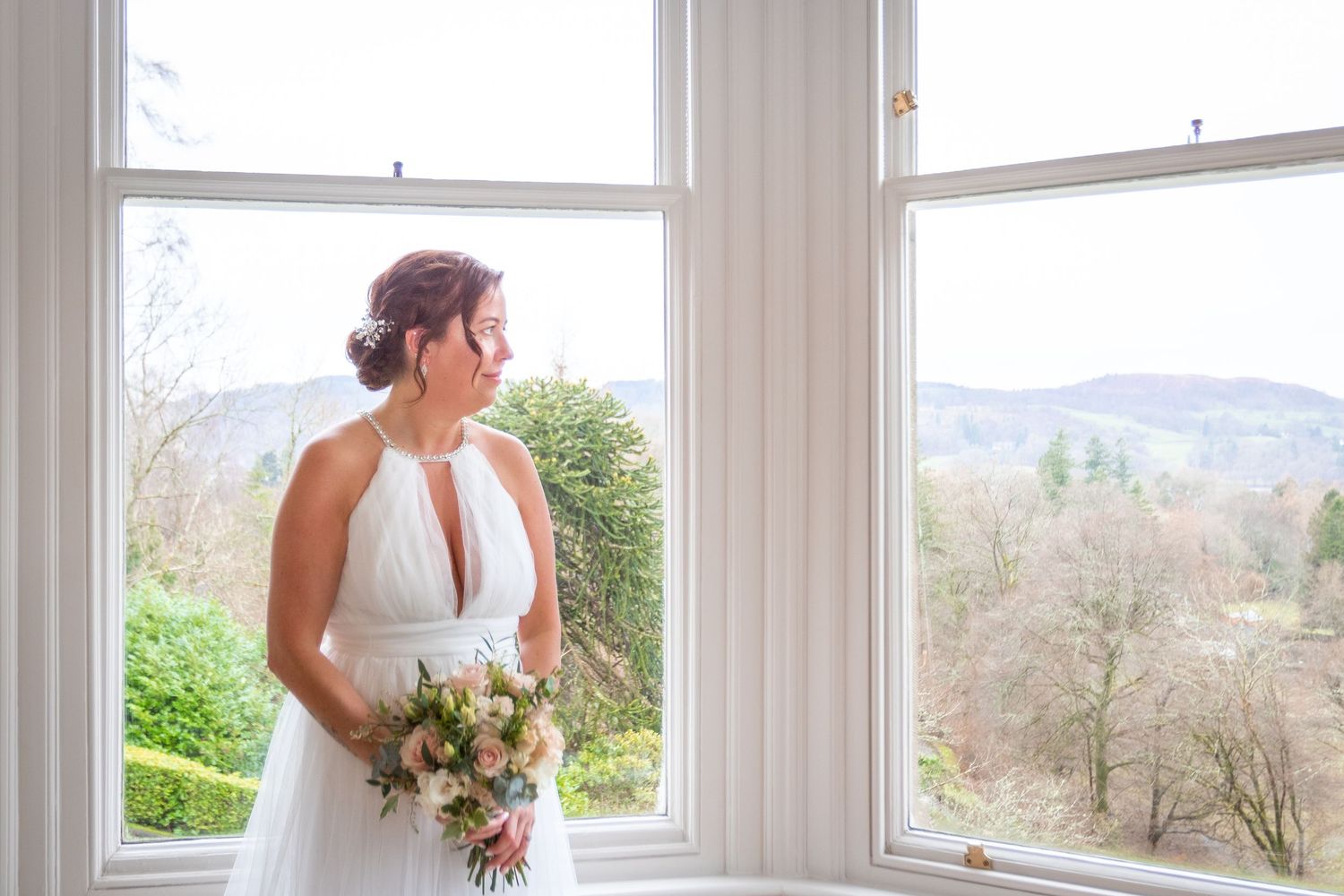 Elegant bride in a halter neck wedding gown holding a bouquet while gazing out a large window.