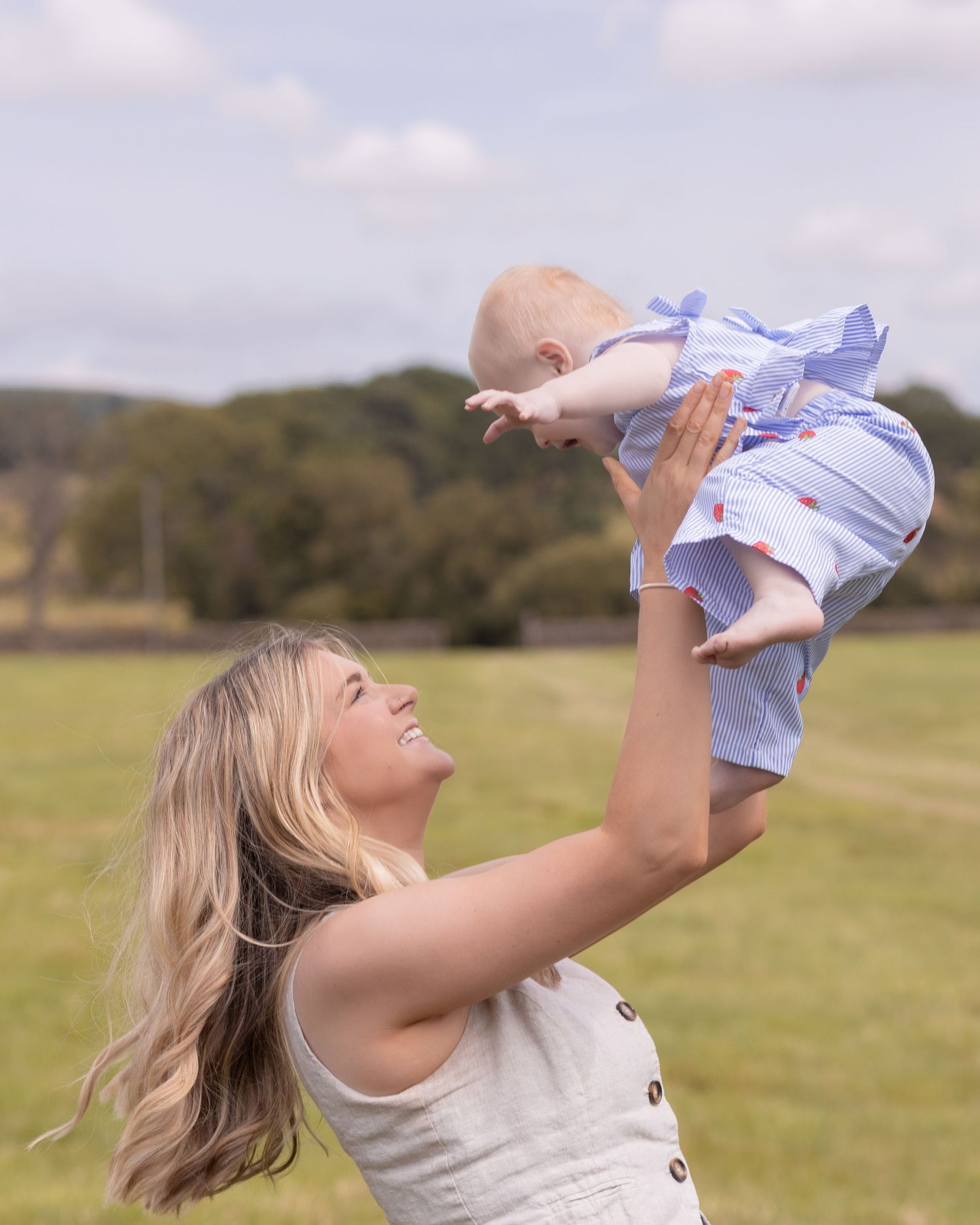 A smiling blonde woman lifts a happy baby in a blue outfit above her head in a sunny green field.