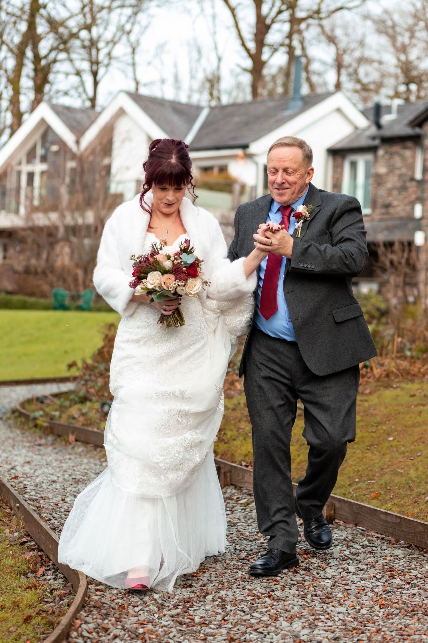Bride in white lace gown holding bouquet walks with suited man in red tie on garden path at wedding venue.