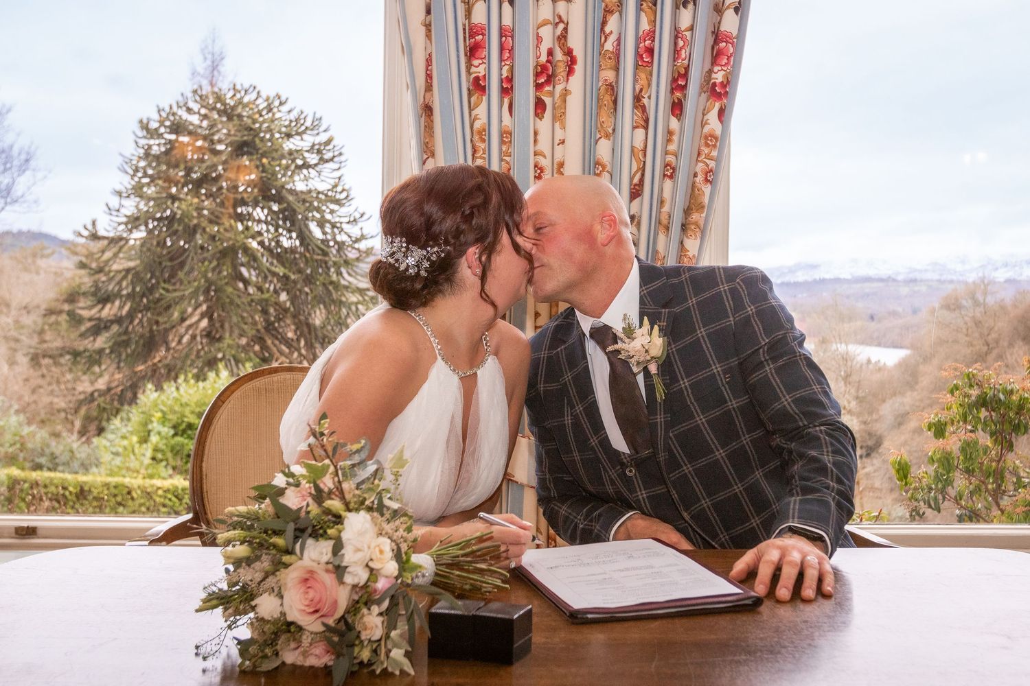 Bride and groom sharing a kiss at the signing table with floral bouquet and scenic window view.