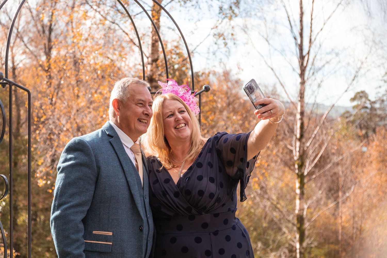Couple taking selfie outdoors under garden arch surrounded by autumn foliage at wedding.