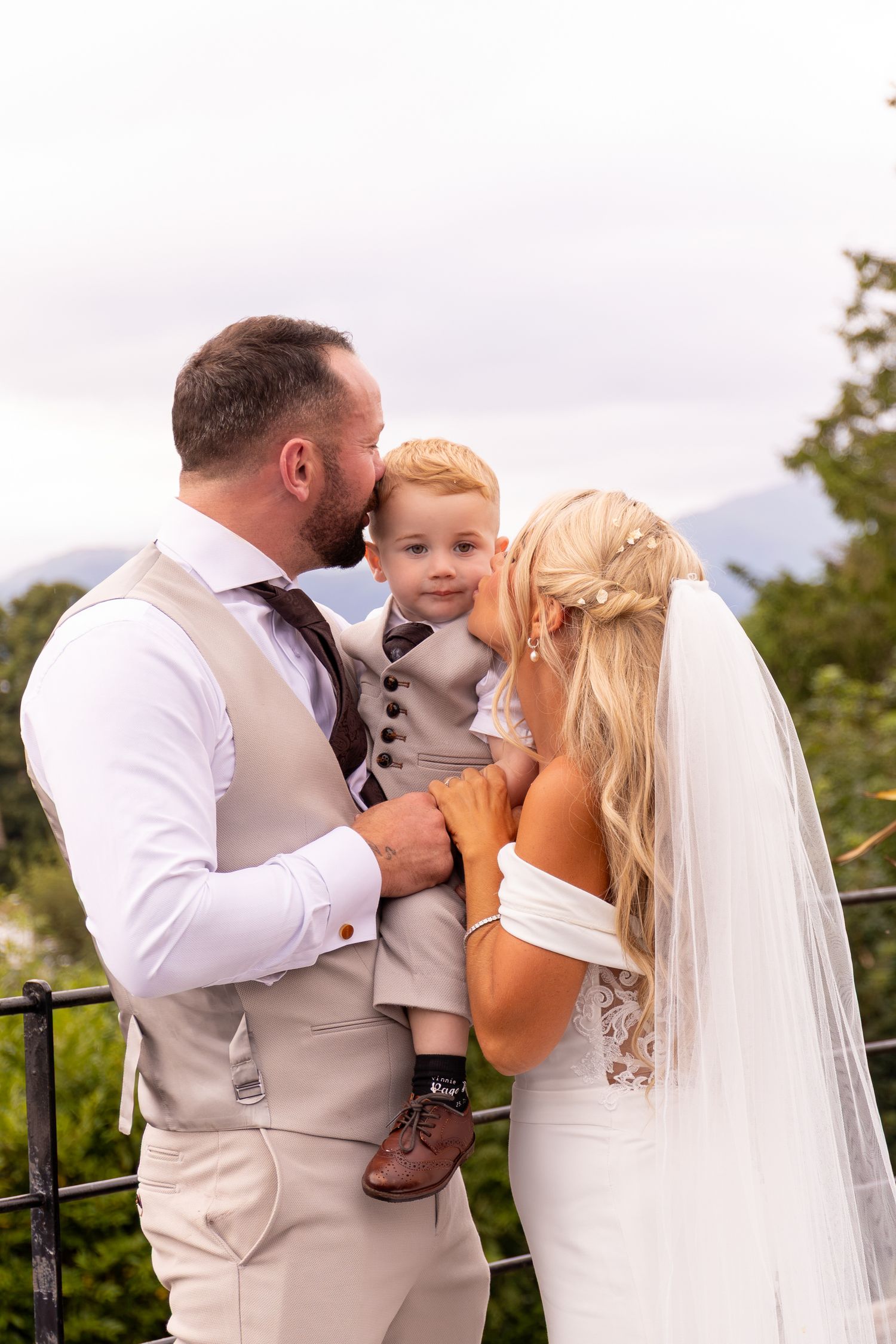 Bride and groom in wedding attire hold young boy in suit, sharing a tender family moment outdoors.