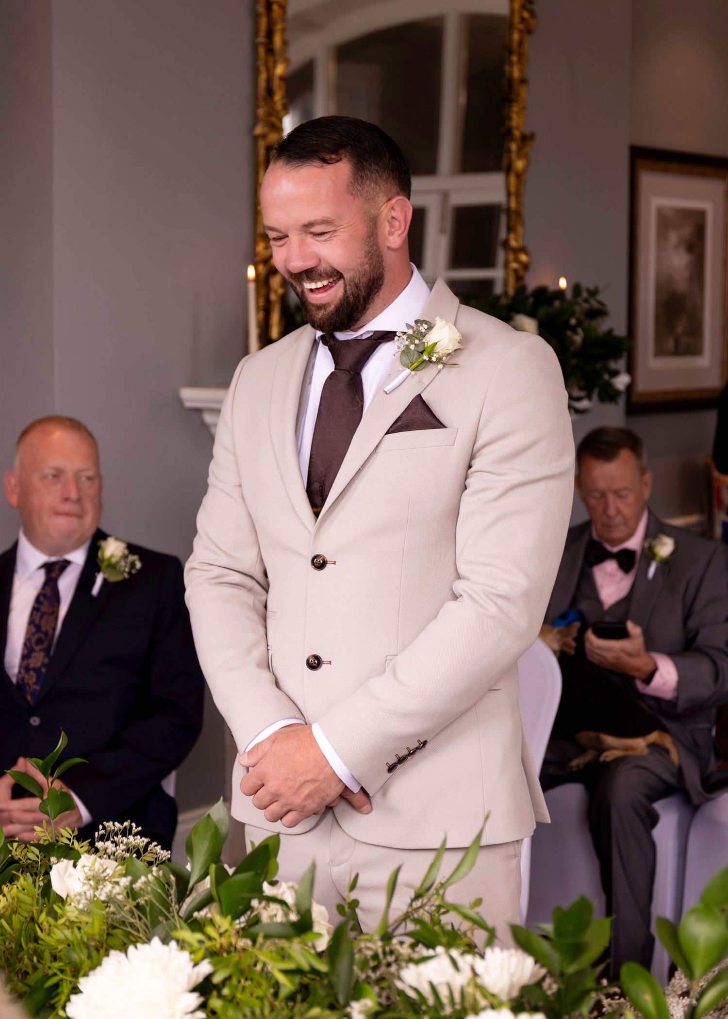 Smiling groom in beige suit with boutonniere stands at wedding altar.