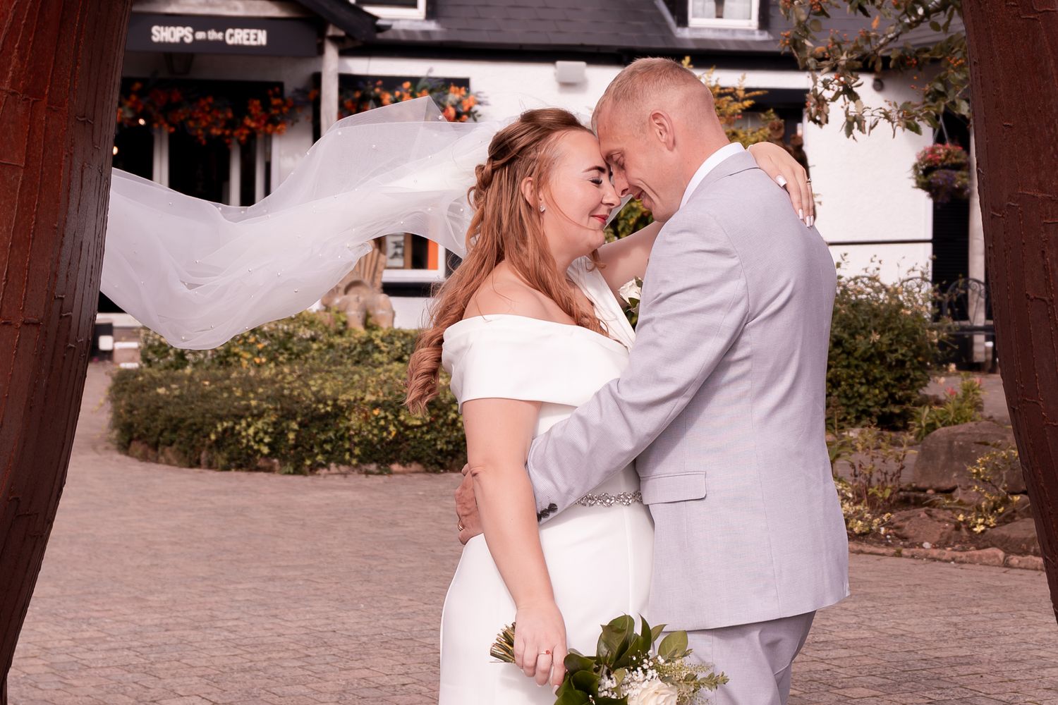 Bride and groom sharing a romantic moment outdoors, her veil flowing in the wind, holding a white bouquet.