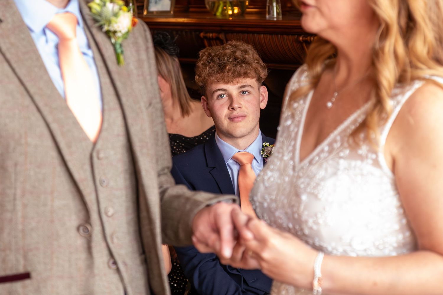 A teenage boy in a navy suit watches closely as a bride and groom hold hands during their wedding.