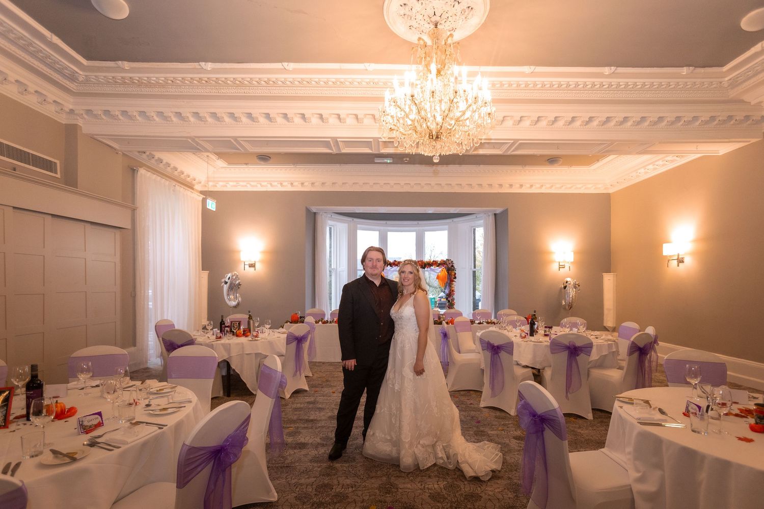 Bride and groom pose in elegant wedding reception hall with chandelier, purple chair sashes, and round tables.