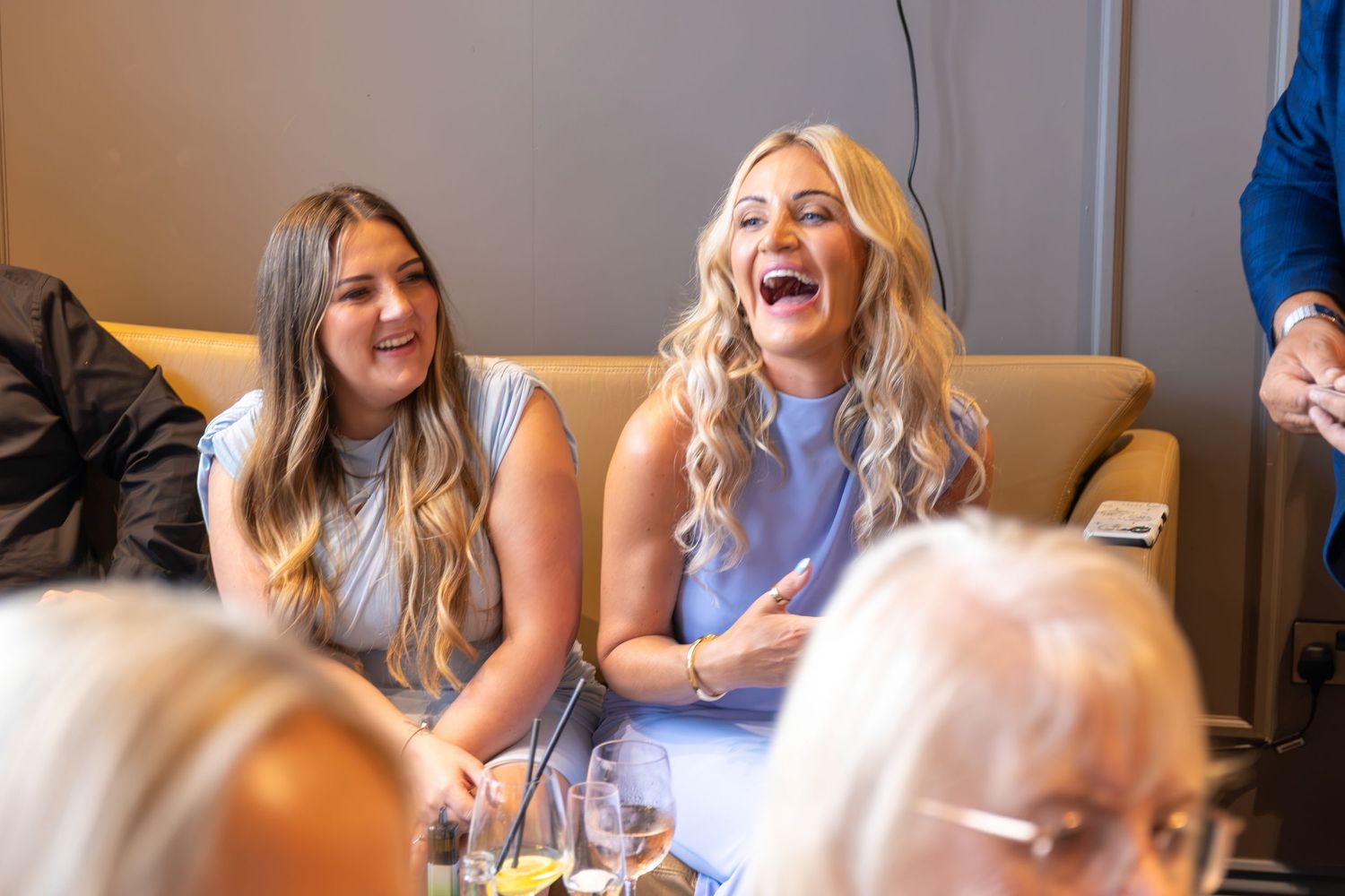 Two women laughing joyfully at an indoor event, wearing light blue outfits, seated in a warmly lit venue.