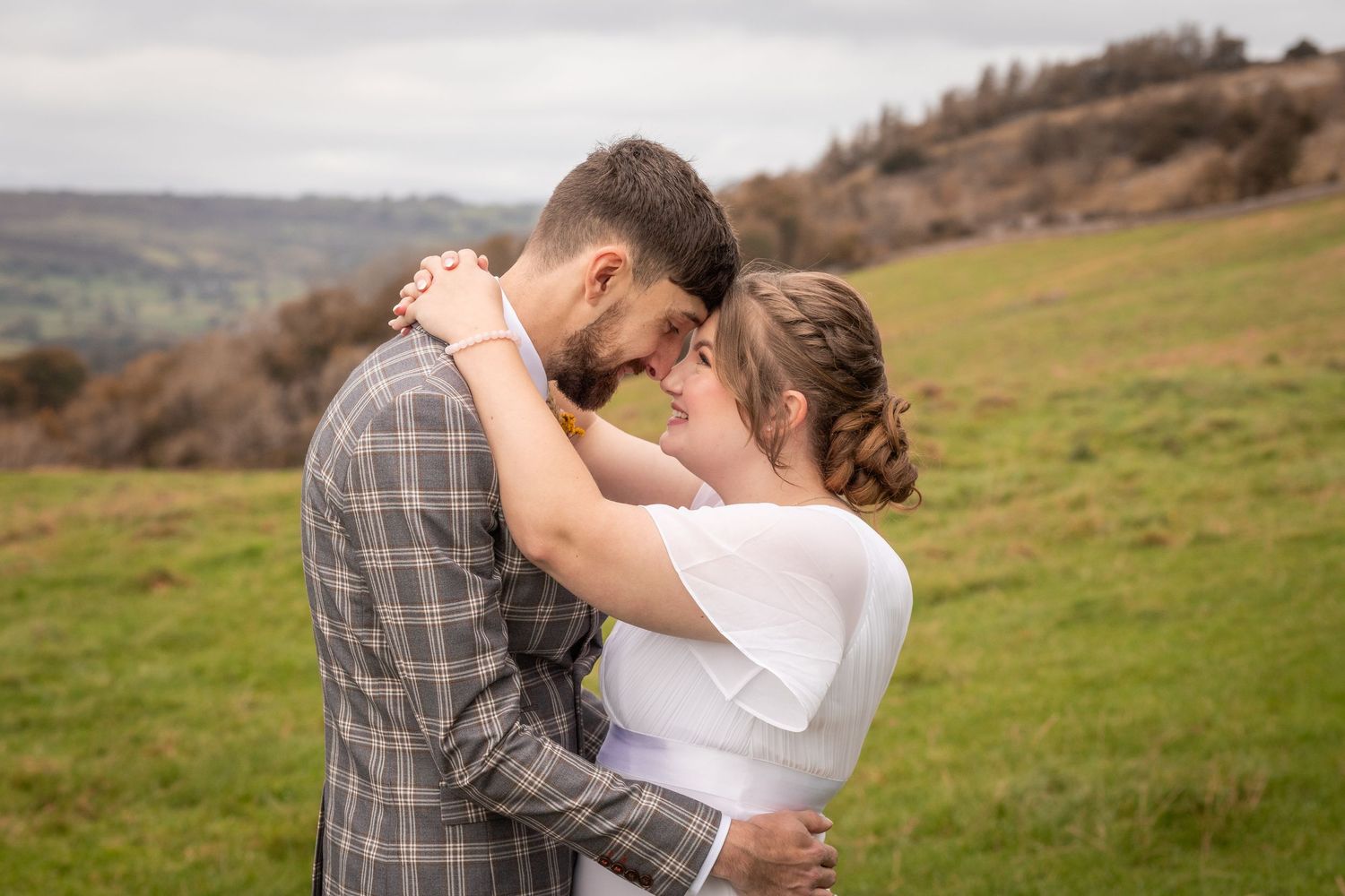 A couple in wedding attire embrace tenderly in a green countryside field, touching foreheads romantically.