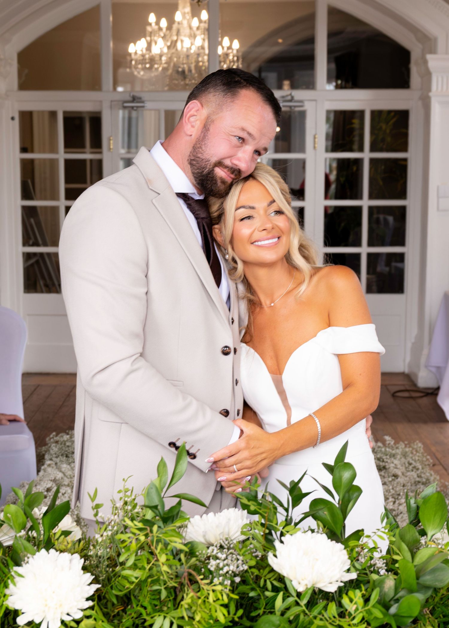 A smiling couple in wedding attire poses together among white floral arrangements in an elegant venue.