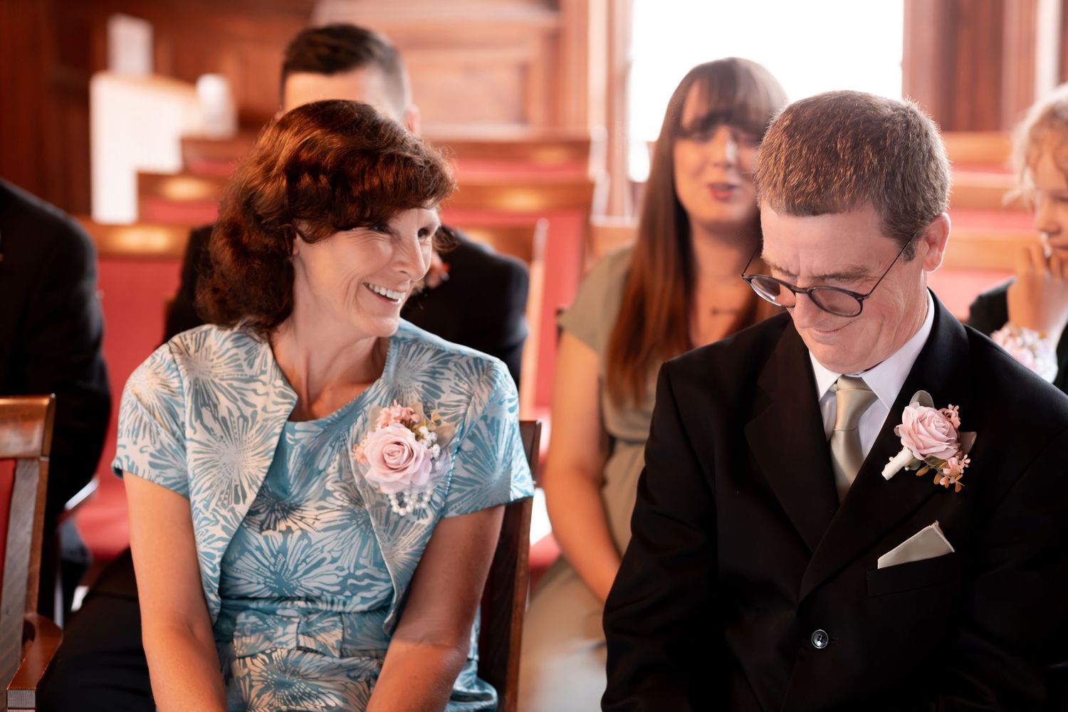 A woman in a blue floral dress and a young man in a suit share a warm moment at a wedding ceremony.