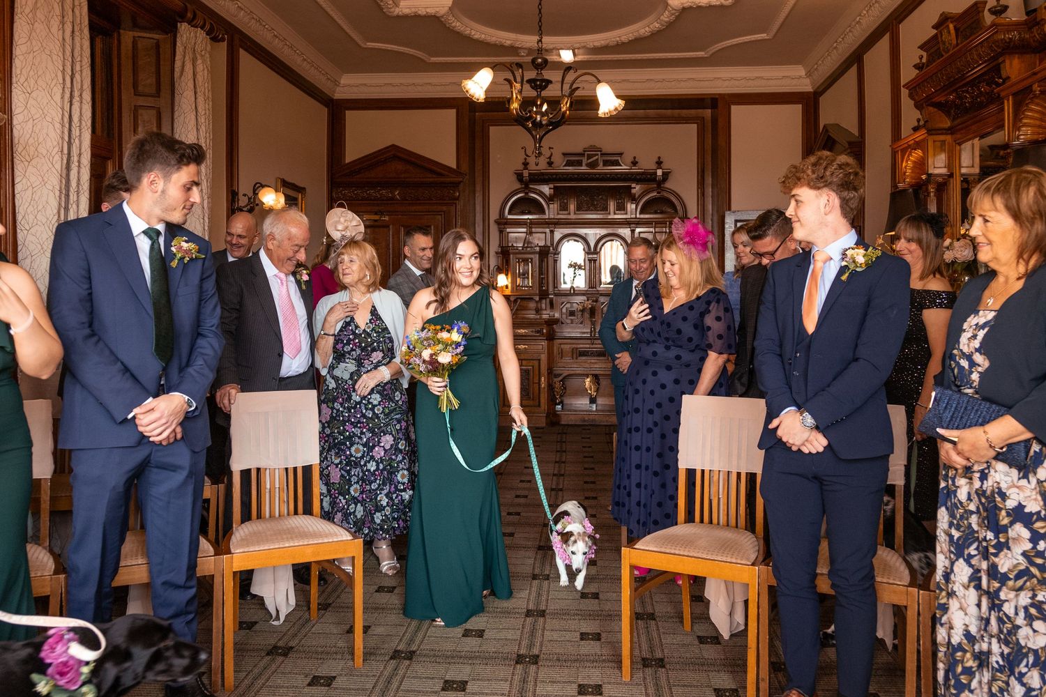 Dog walks bride down the aisle at elegant wedding ceremony with guests standing in ornate venue.