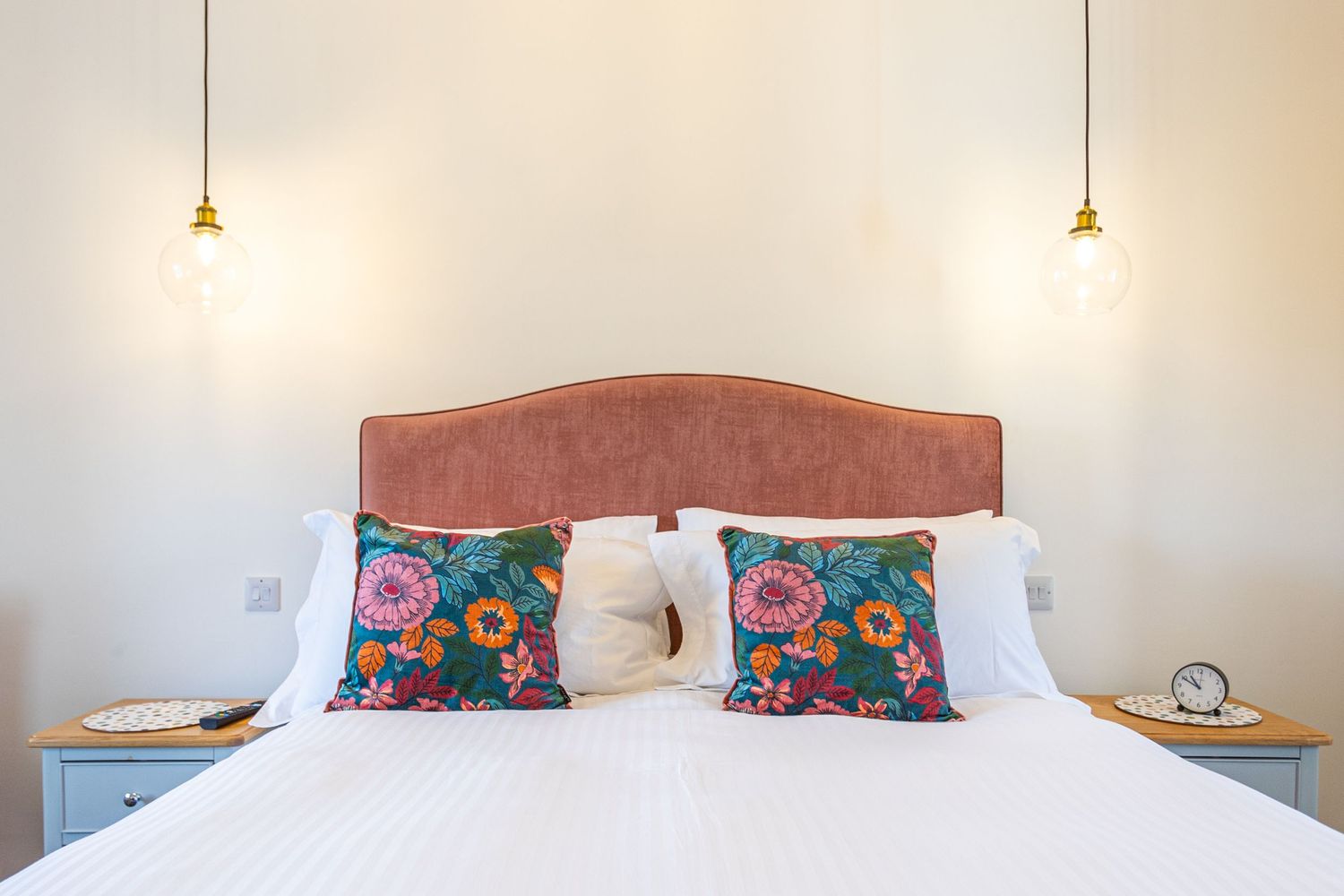 Cozy bedroom with pink velvet headboard, floral cushions, and warm Edison bulb pendant lights.