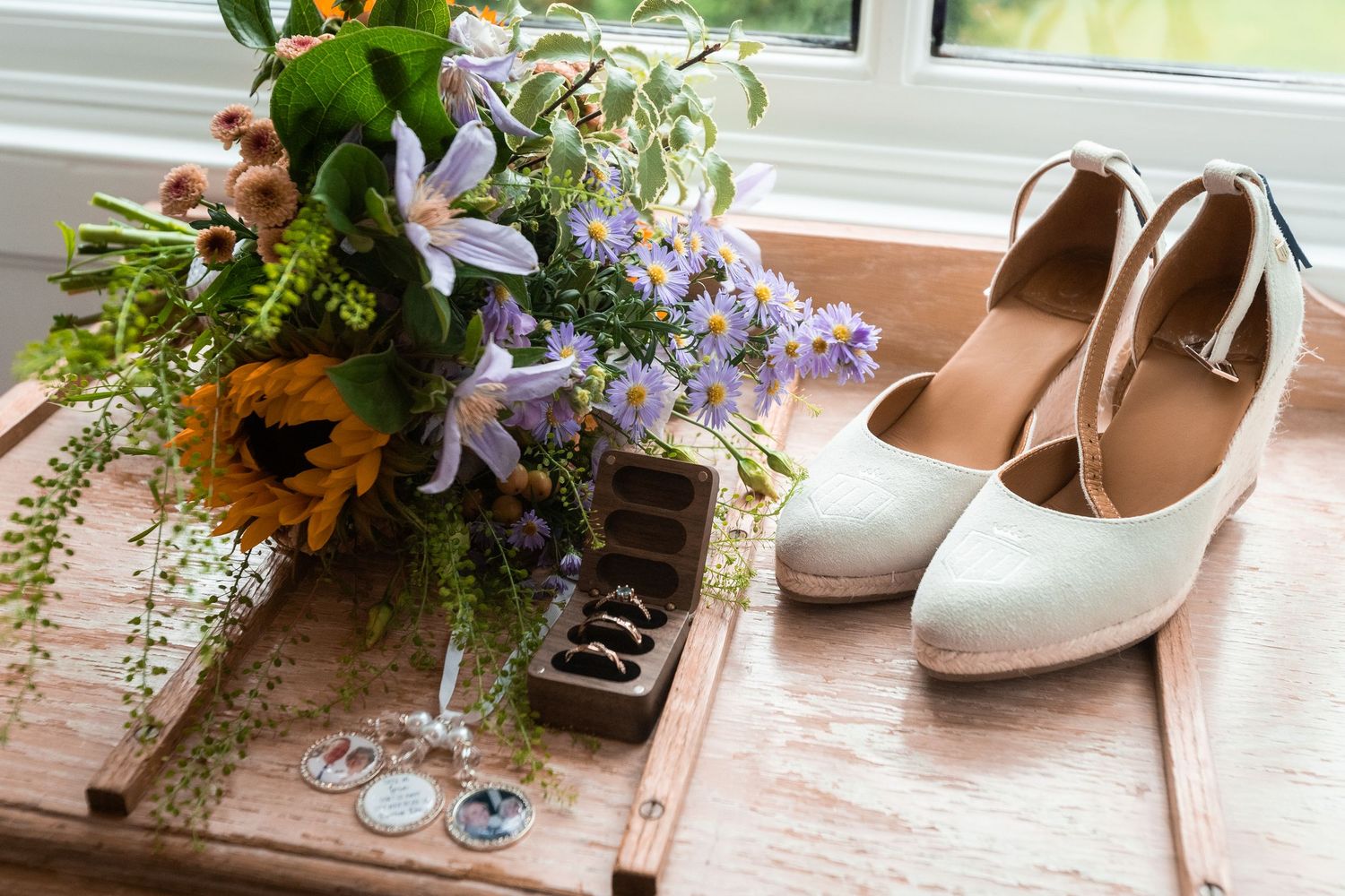 White bridal heels beside a wildflower bouquet, ring box, and earrings on a rustic wooden surface near a window.